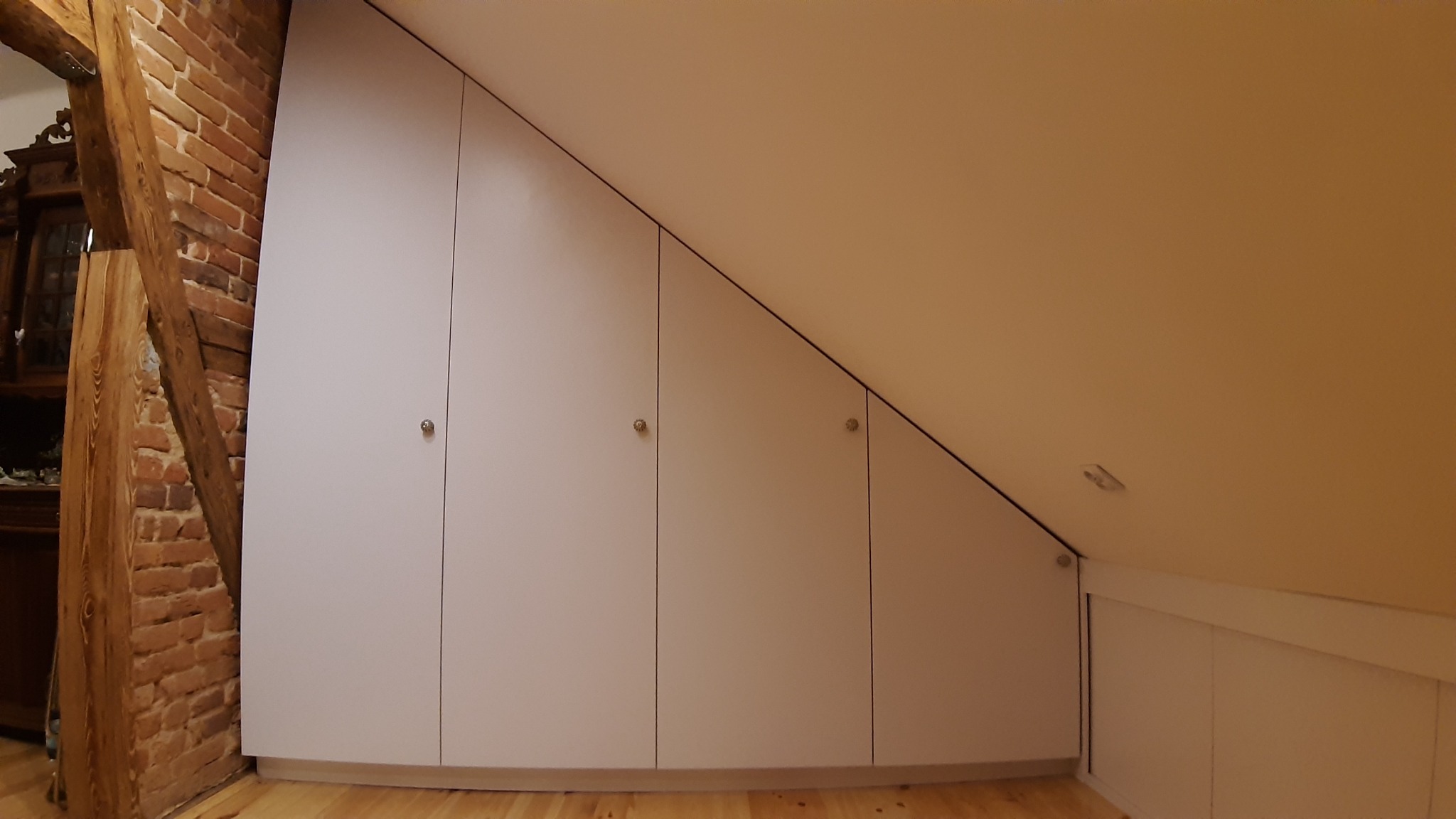 Biała szafa wnękowa z czterema drzwiami, dopasowana do skosu poddasza, z okrągłymi gałkami, na drewnianej podłodze, obok fragmentu ściany z cegły.
