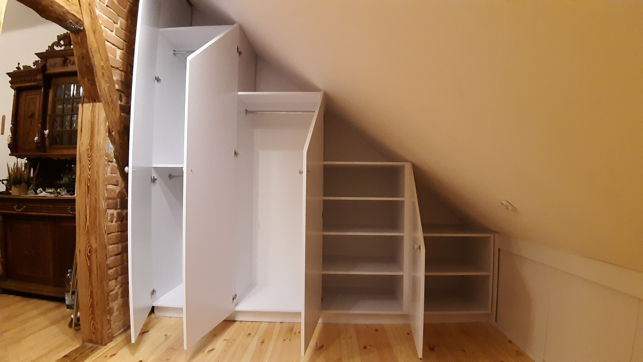 Biała zabudowa szafy wnękowej pod skosem na poddaszu, widoczne półki i drążek, otwarte drzwi, drewniana podłoga i fragment ściany z cegły w tle.