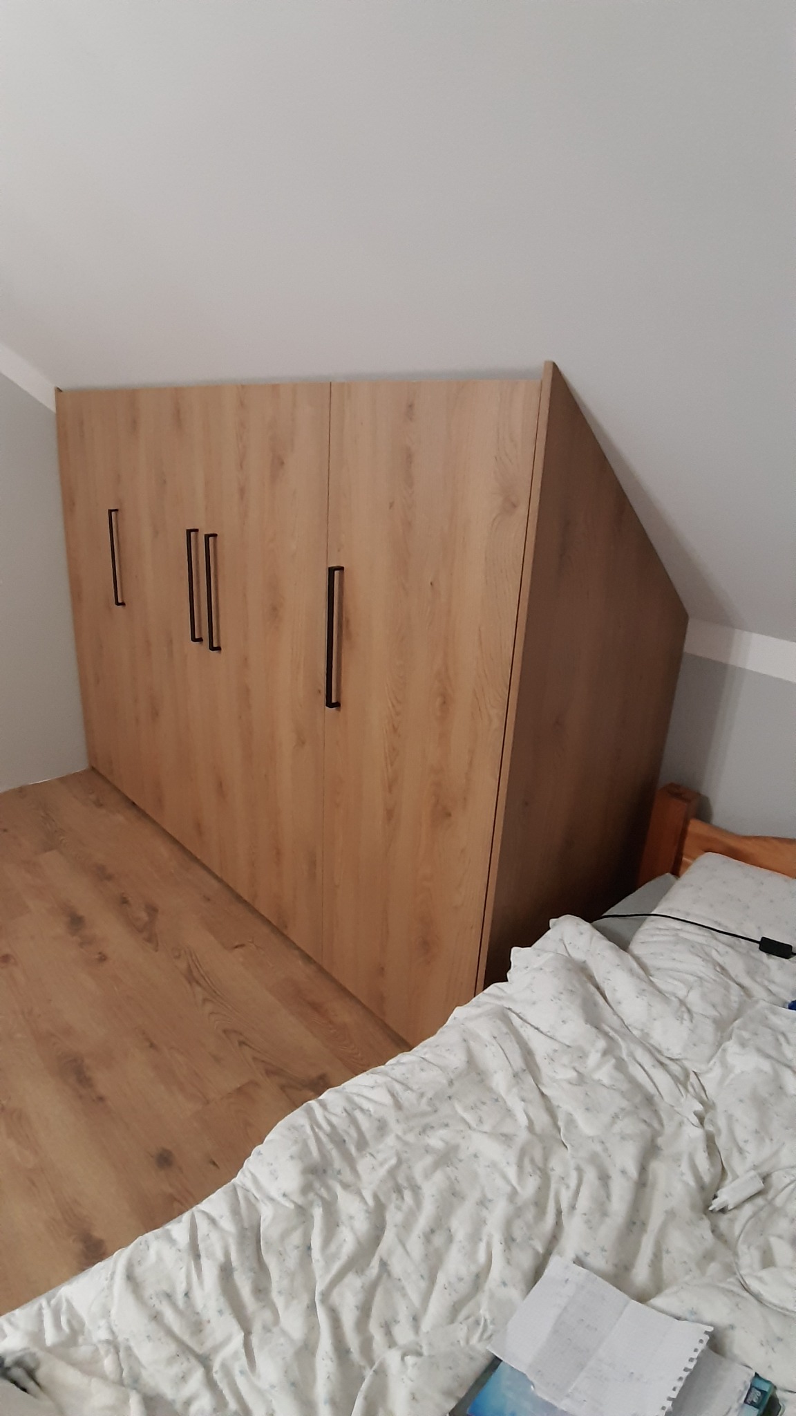 Trzydrzwiowa, jasna szafa z ciemnymi uchwytami, dopasowana do skosu dachu w pokoju, widoczna częściowo pościel na łóżku.