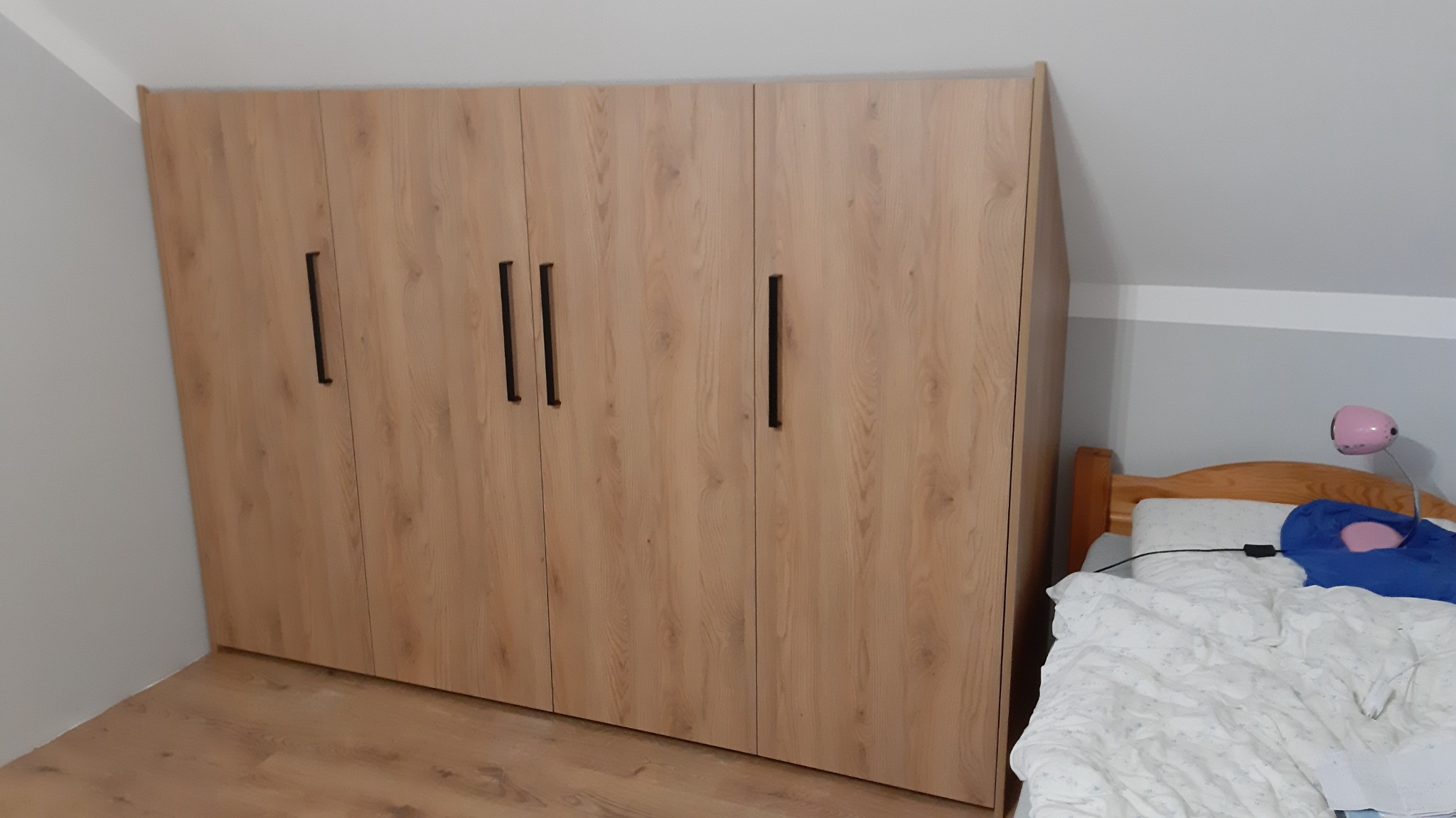 Duża szafa z frontami w kolorze jasnego drewna z czarnymi, pionowymi uchwytami, dopasowana do skosu dachu w pokoju z drewnianą podłogą i łóżkiem w tle.