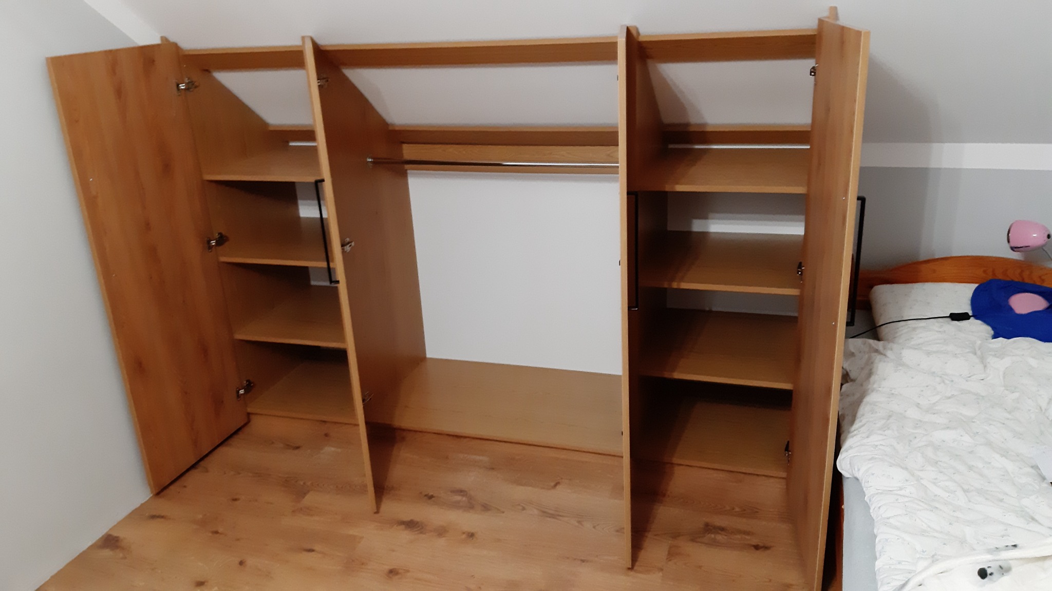 Otwarta, drewniana szafa wnękowa z półkami i drążkiem na ubrania, widoczna w sypialni z łóżkiem w tle.