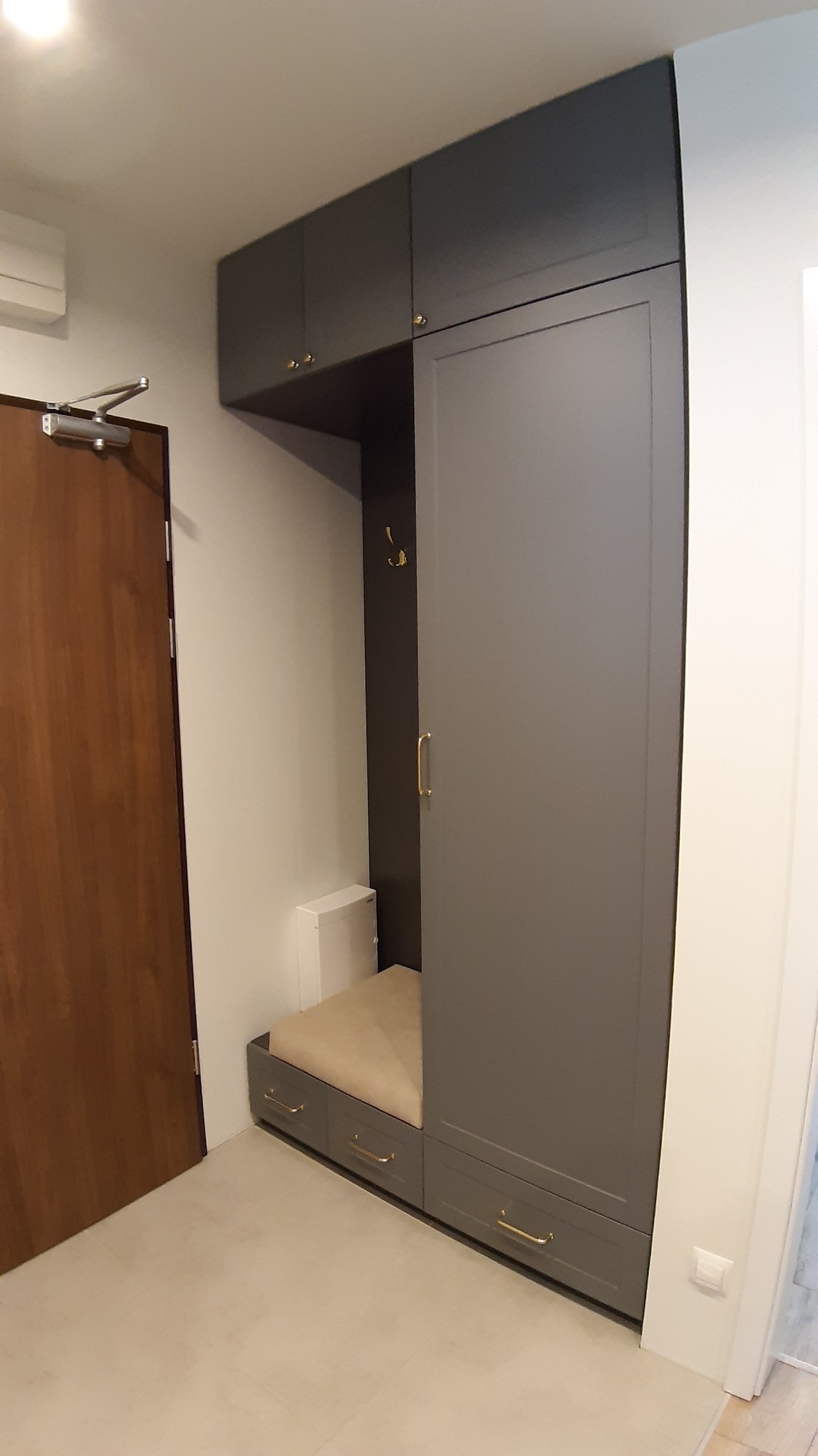 Szara szafa wnękowa z siedziskiem i złotymi uchwytami, zamontowana w przedpokoju obok brązowych drzwi.