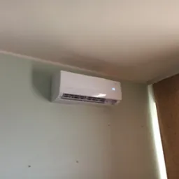 Biała klimatyzacja ścienna z wyświetlaczem temperatury 26 stopni Celsjusza, zamontowana na jasnej, zielonkawej ścianie w pomieszczeniu z zasłoną w tle.