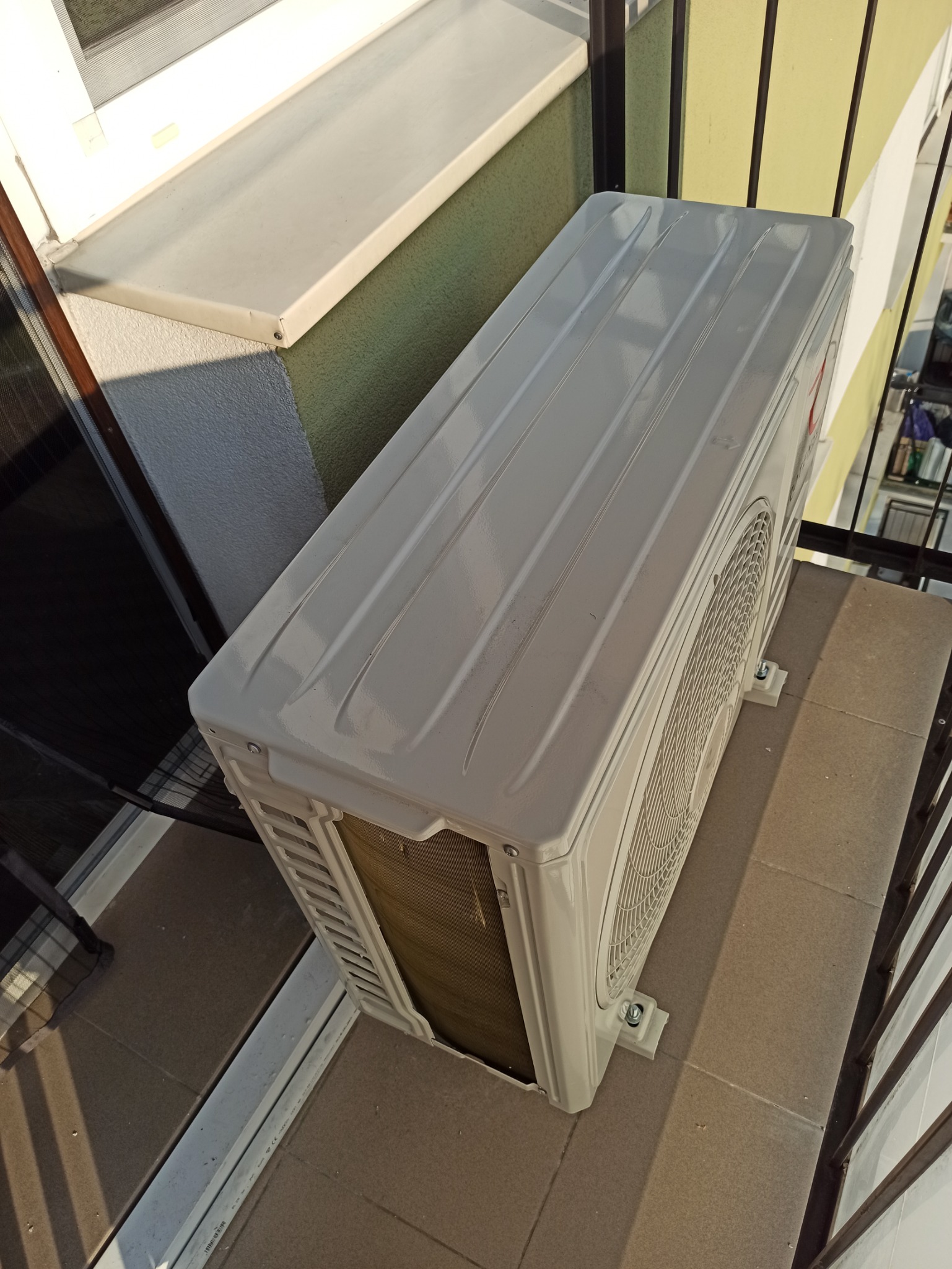 Zewnętrzna jednostka klimatyzacji zamontowana na balkonie, widoczna z góry, z metalową kratką wentylacyjną i elementami mocującymi do podłoża.