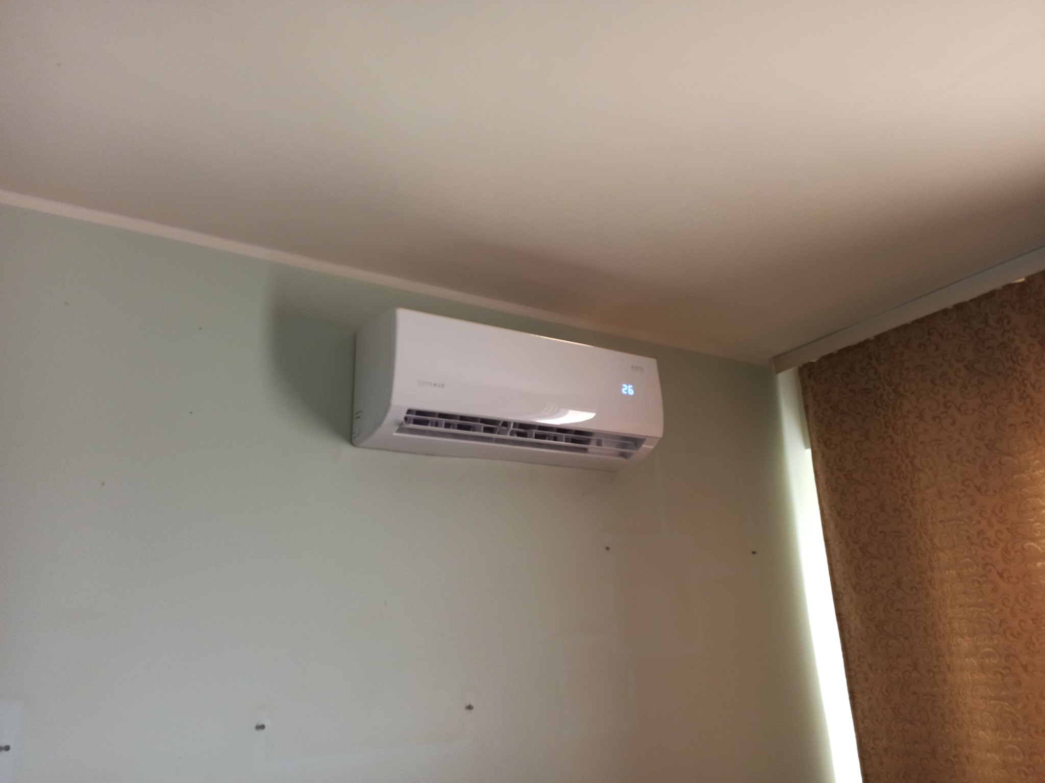 Biała klimatyzacja ścienna z wyświetlaczem temperatury 26 stopni Celsjusza, zamontowana na jasnej, zielonkawej ścianie w pomieszczeniu z zasłoną w tle.