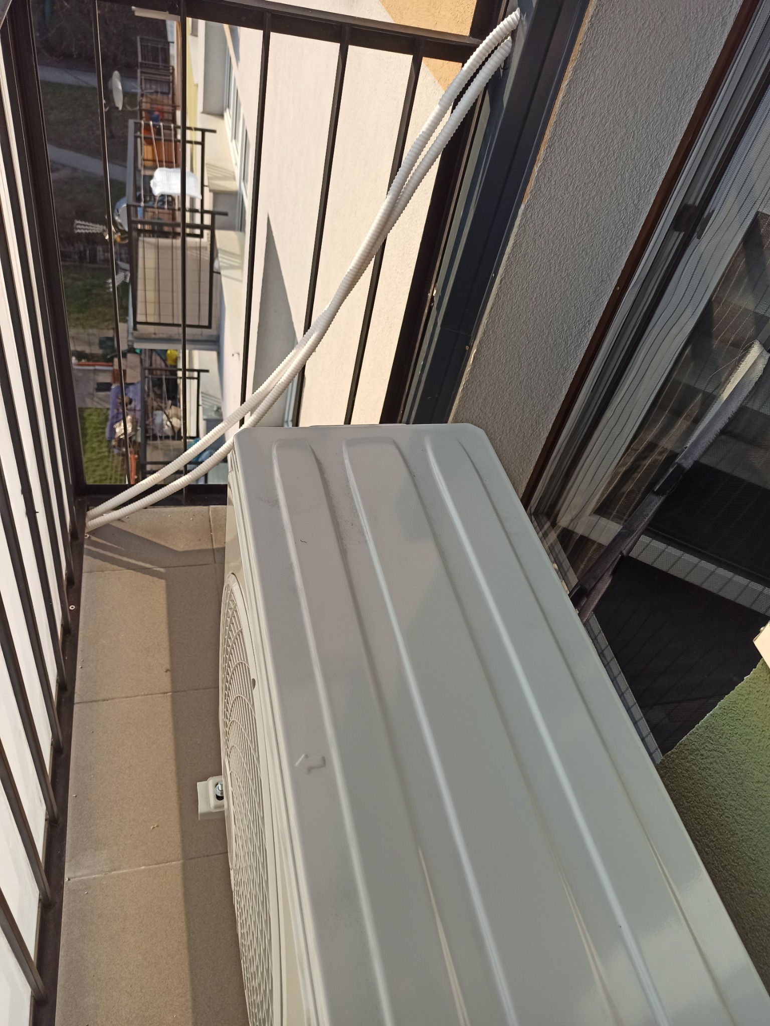 Zewnętrzna jednostka klimatyzacji zamontowana na balkonie, widoczne przewody prowadzące do wnętrza budynku, balkon z metalową balustradą.