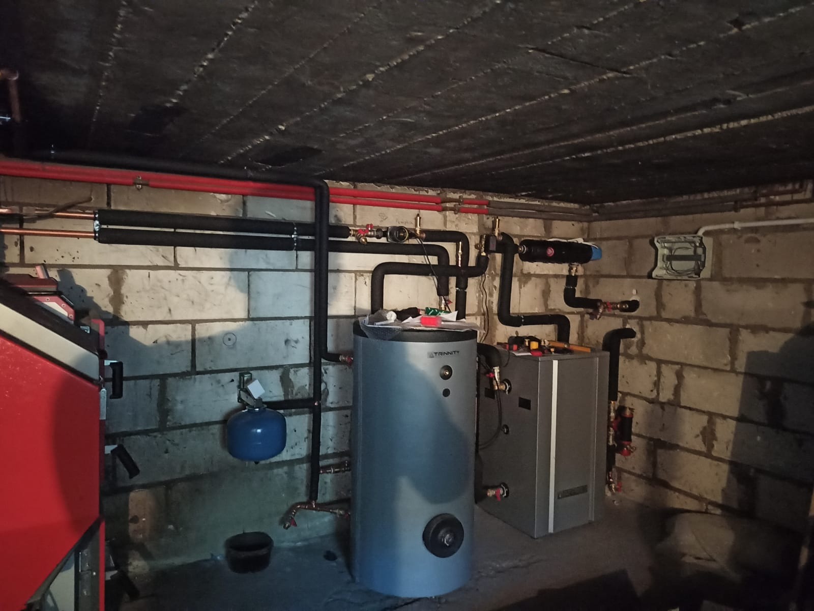 Zestaw urządzeń grzewczych w kotłowni: srebrny zasobnik ciepłej wody marki Trinity, obok szary piec, widoczne rury miedziane i czarne izolowane, na ścianie z cegły instalacja elektryczna.