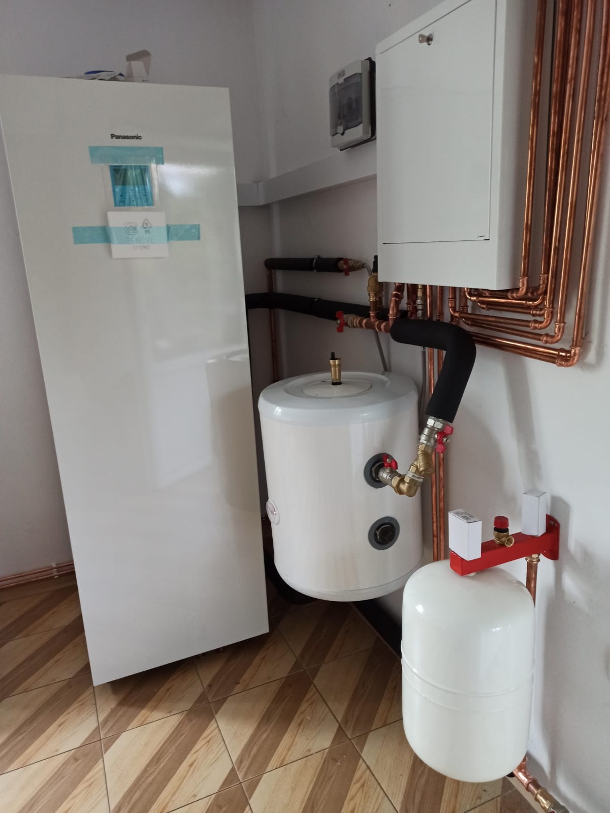 Biała pompa ciepła Panasonic z zbiornikiem buforowym i instalacją z miedzianych rur, widoczne przyłącza i zawory, na podłodze brązowe płytki.