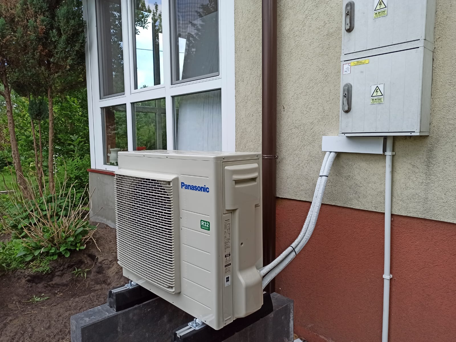 Zewnętrzna jednostka klimatyzacji Panasonic R32 zamontowana na betonowym fundamencie przy ścianie domu z brązowym orynnowaniem i skrzynką elektryczną na elewacji.