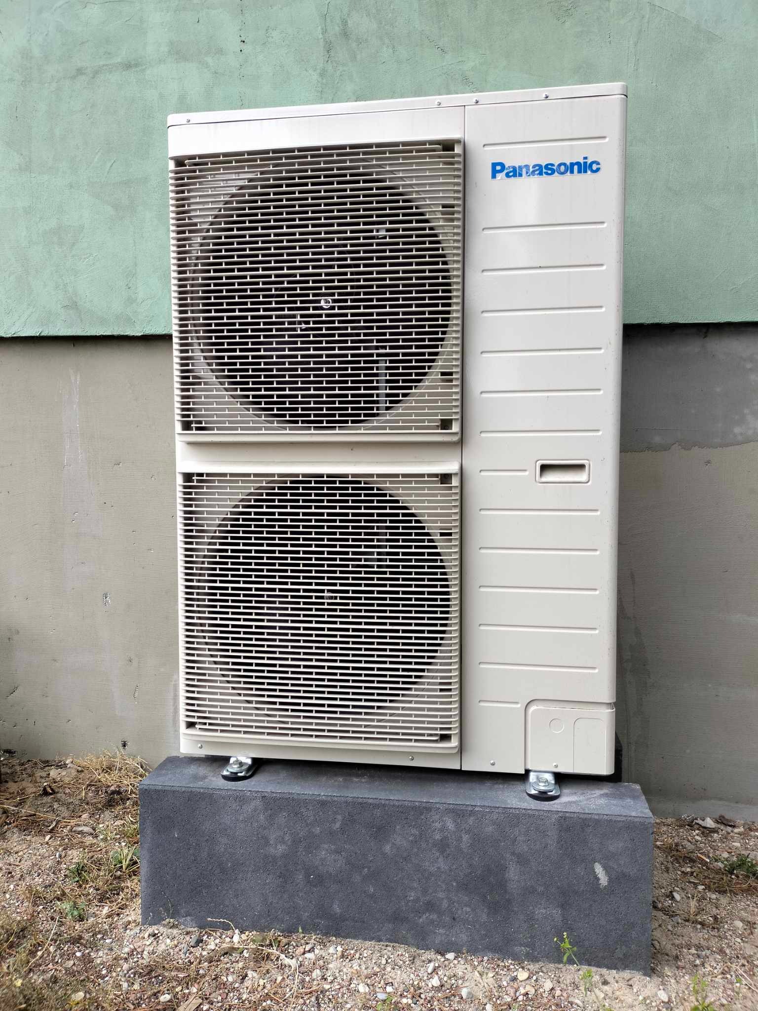 Zewnętrzna jednostka klimatyzacji Panasonic, zamontowana na betonowym cokole przy ścianie budynku o zielonkawej elewacji.
