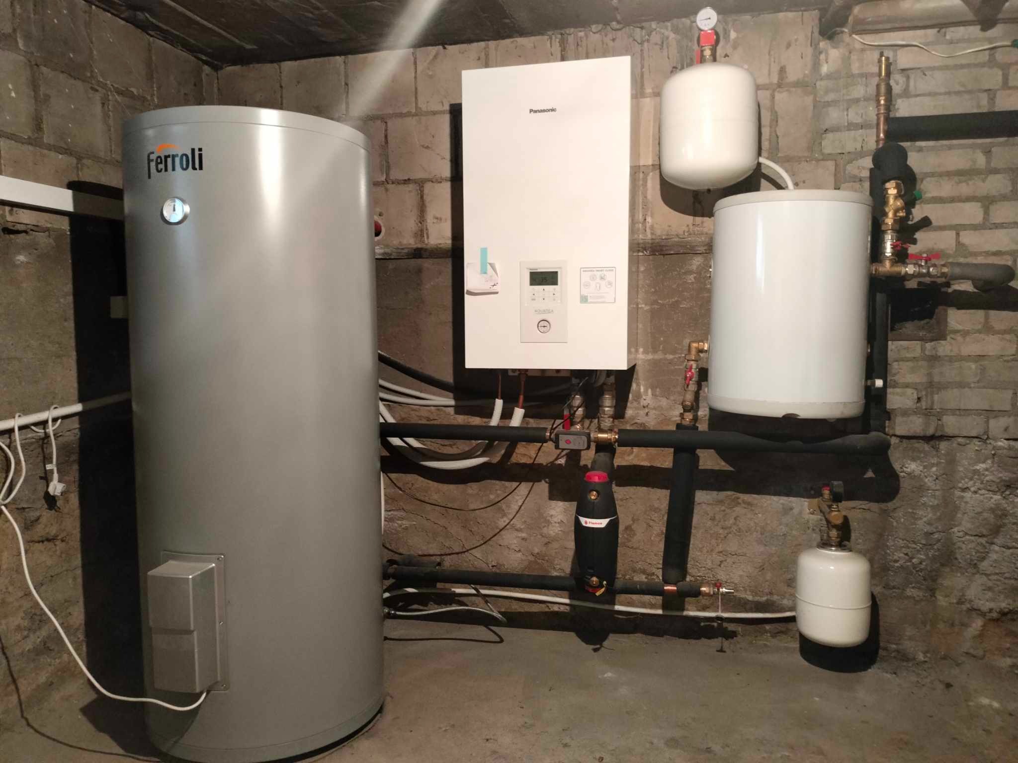 Instalacja pompy ciepła Panasonic Aquarea z zasobnikiem Ferroli w piwnicy o ścianach z cegły, widoczne rury i zbiorniki wyrównawcze.