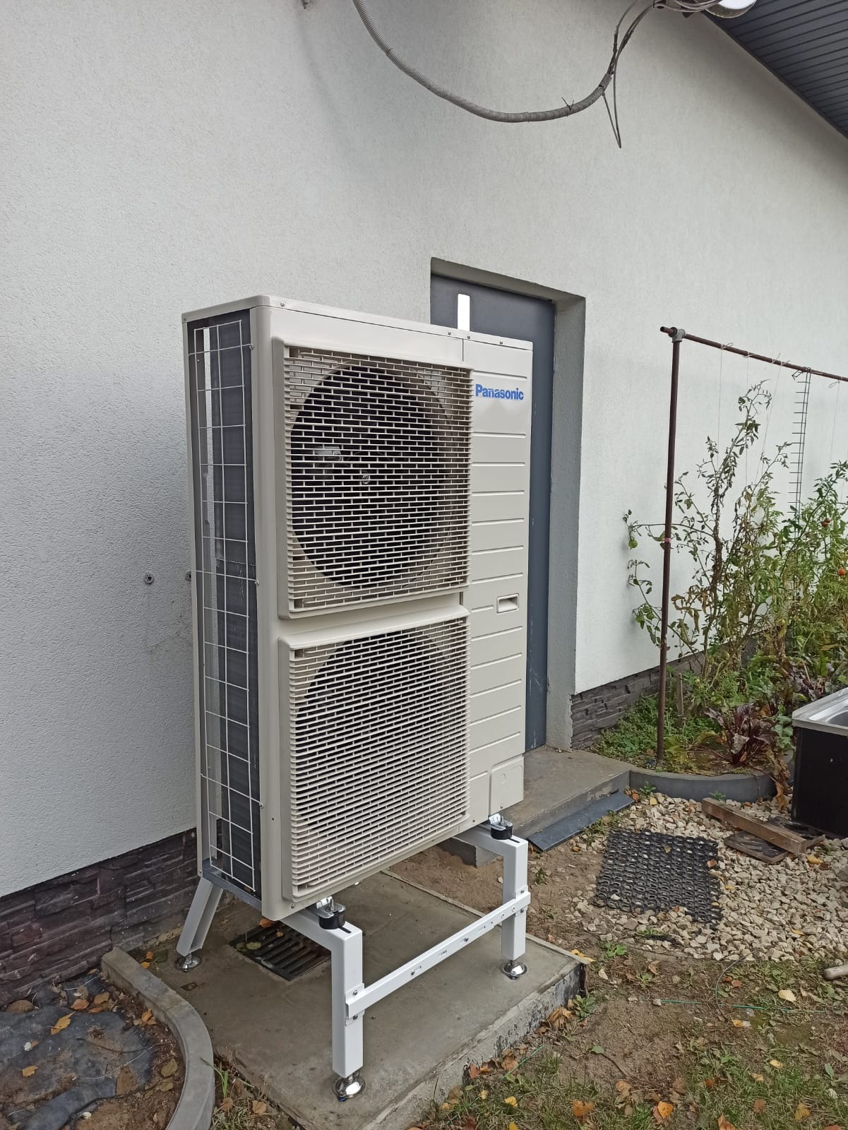 Zewnętrzna jednostka pompy ciepła Panasonic, zamontowana na białym, metalowym stelażu, na betonowej podstawie, obok ściany budynku z widocznymi elementami instalacji elektrycznej i roślinnością w tle.