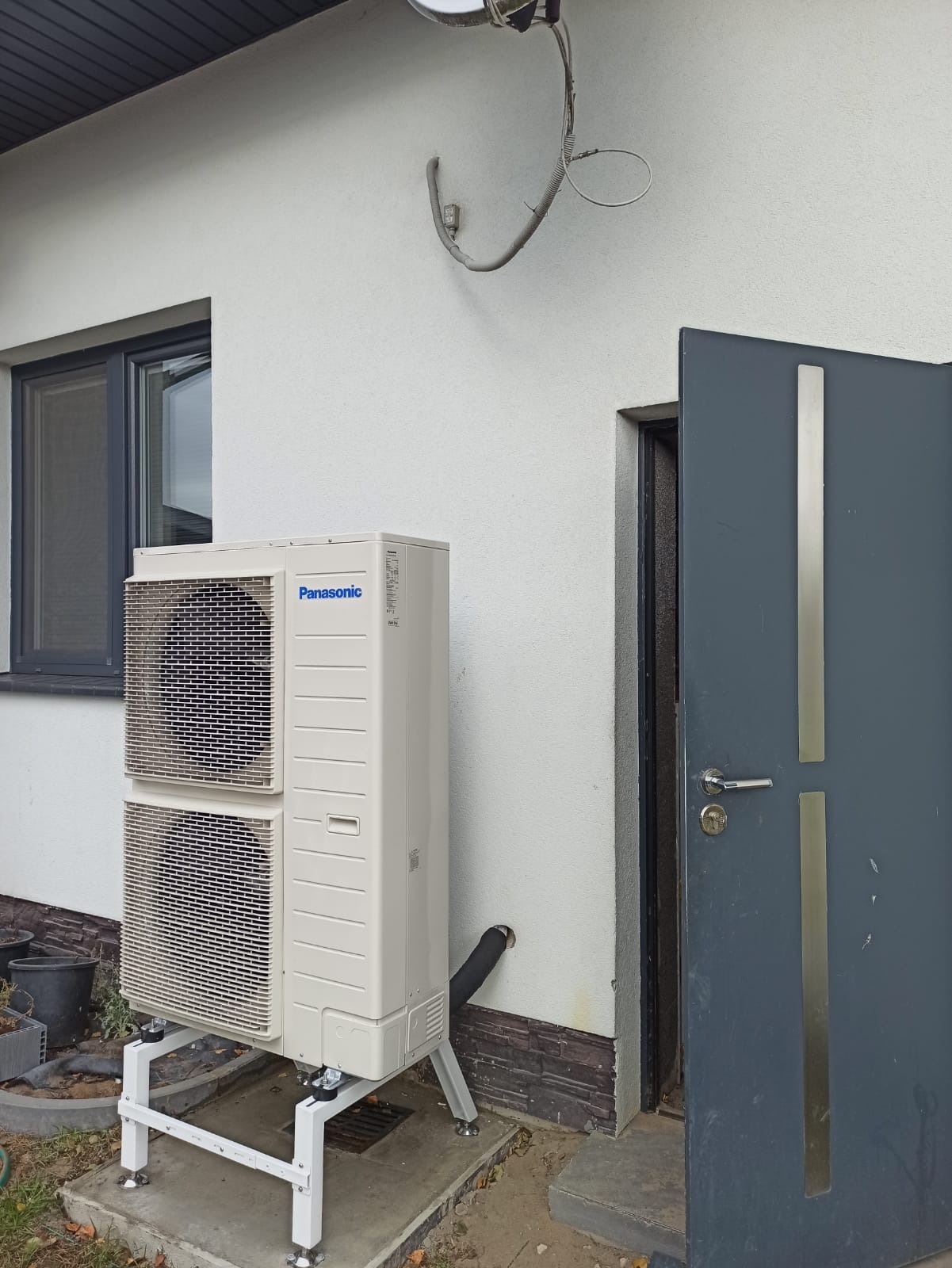 Biała pompa ciepła Panasonic na metalowym stelażu umieszczona obok szarych drzwi wejściowych i okna w jasnym budynku z zewnętrzną instalacją kablową.