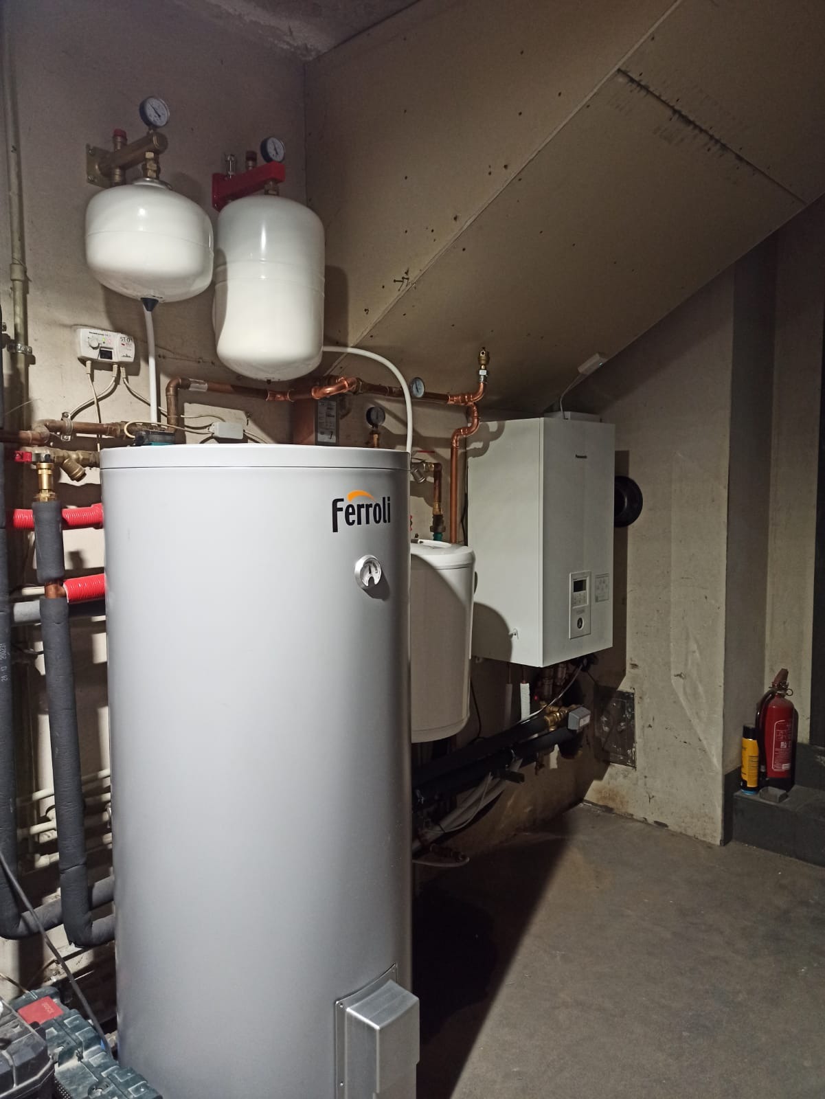 Szary bojler Ferroli z podłączonymi rurami miedzianymi, obok biały piec grzewczy na tle betonowej ściany w piwnicy, widoczna gaśnica i narzędzia.