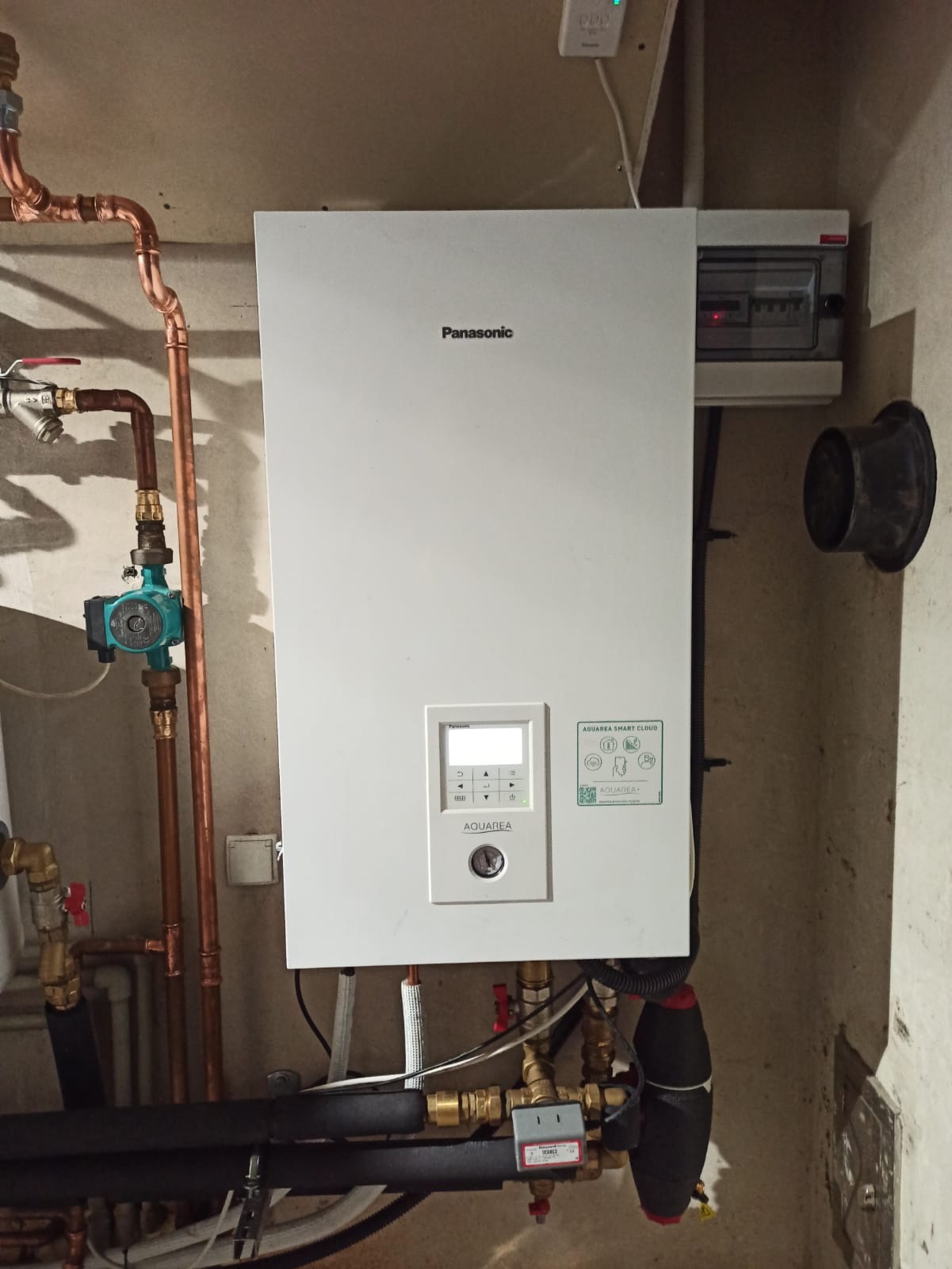 Biała pompa ciepła Panasonic Aquarea Smart Cloud zamontowana na ścianie, widoczne miedziane rury, zawory i czarna izolacja termiczna rur. Obok skrzynka elektryczna i otwór wentylacyjny.