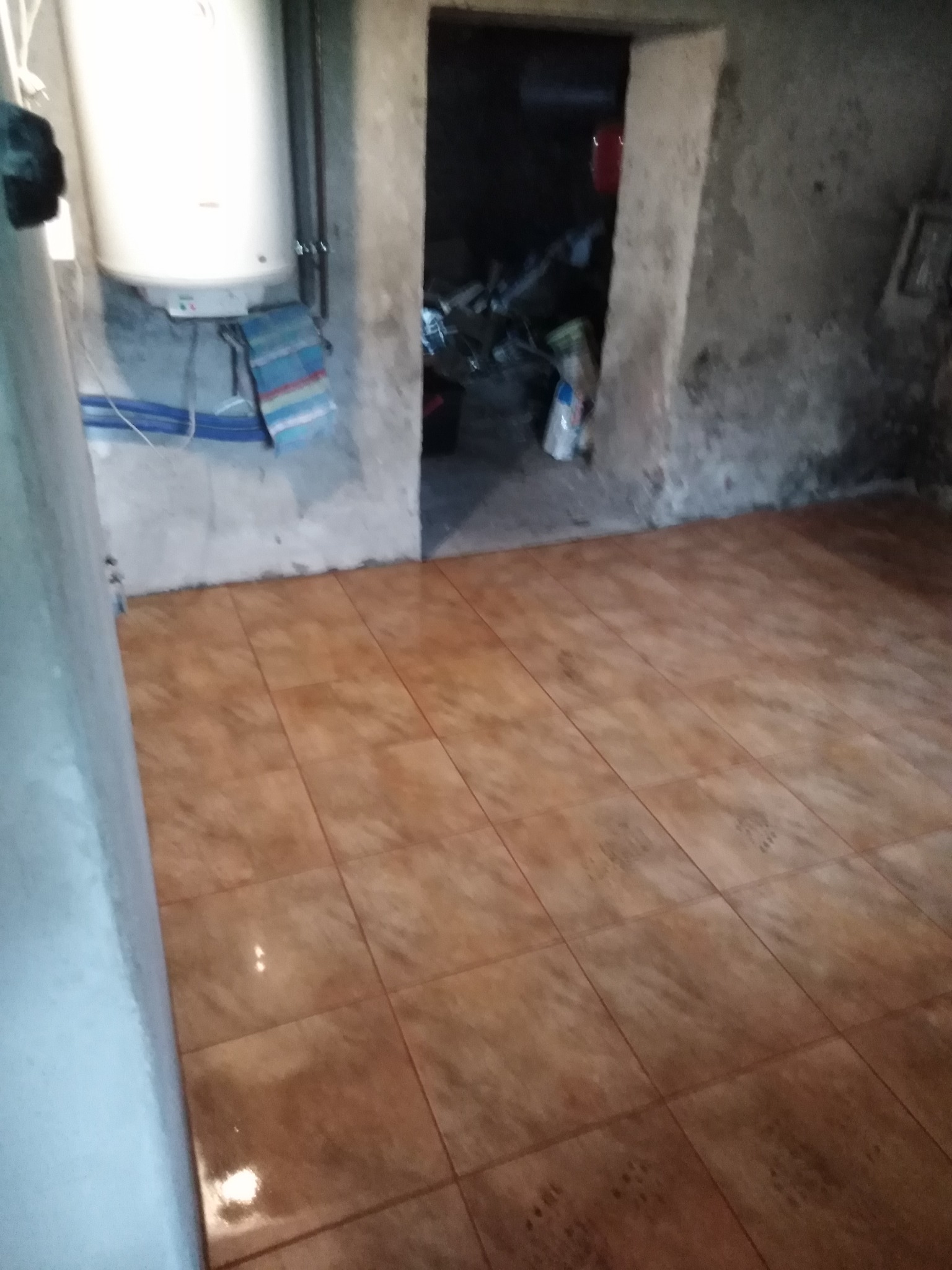 Piwnica z nową, brązową podłogą z płytek ceramicznych; na ścianie biały bojler, a w tle otwarte drzwi do ciemnego pomieszczenia.