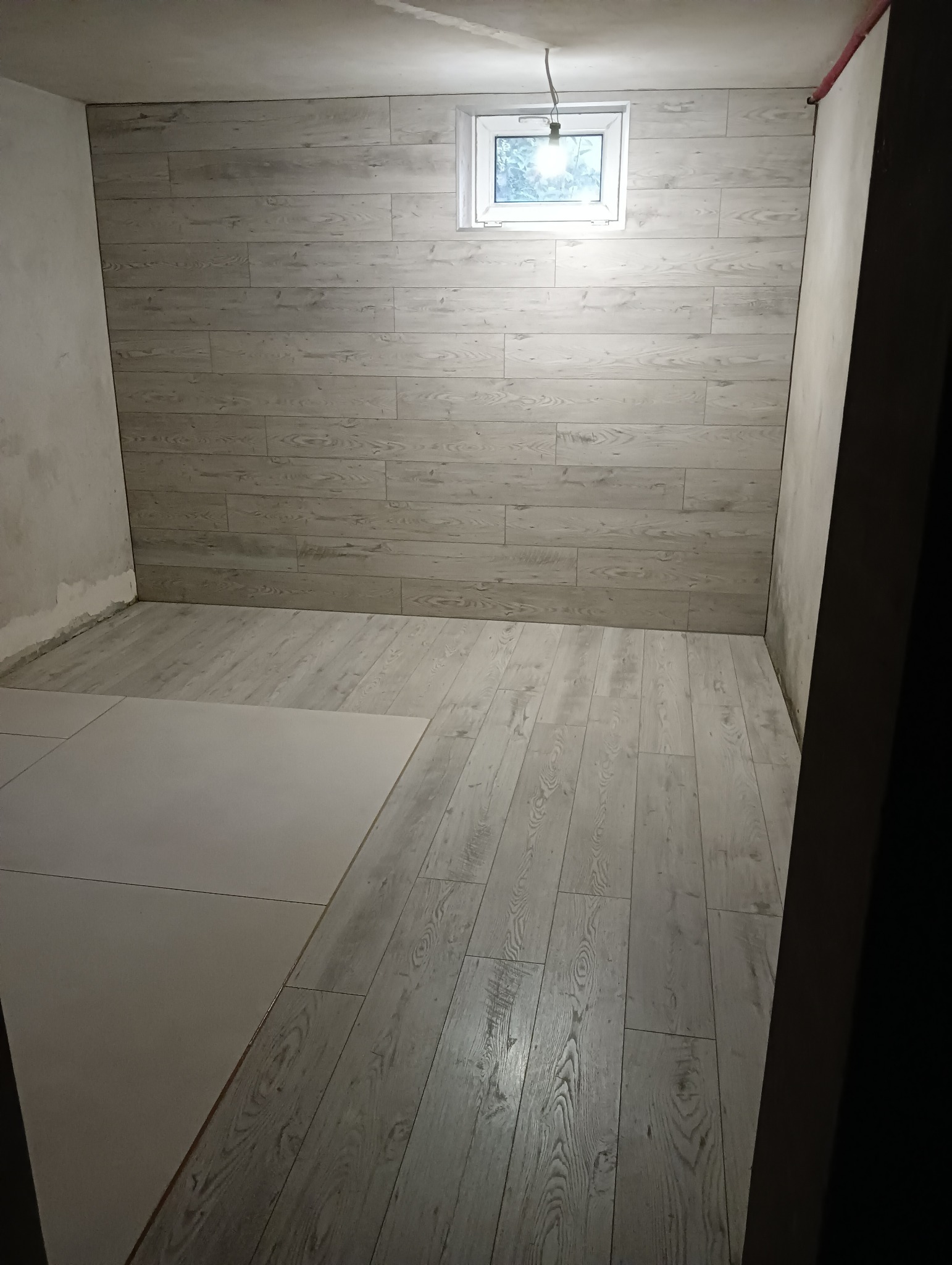 Wnętrze pokoju z wykończonymi ścianami panelami imitującymi drewno i częściowo ułożoną podłogą z paneli, widoczne okno z widokiem na zieleń.