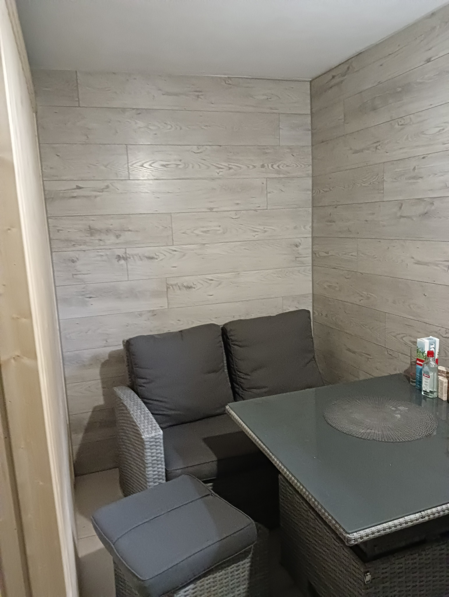 Zabudowany balkon z szarą sofą rattanową, pufem i szklanym stolikiem, ściany wykończone panelami drewnopodobnymi, widoczne butelki na stole.