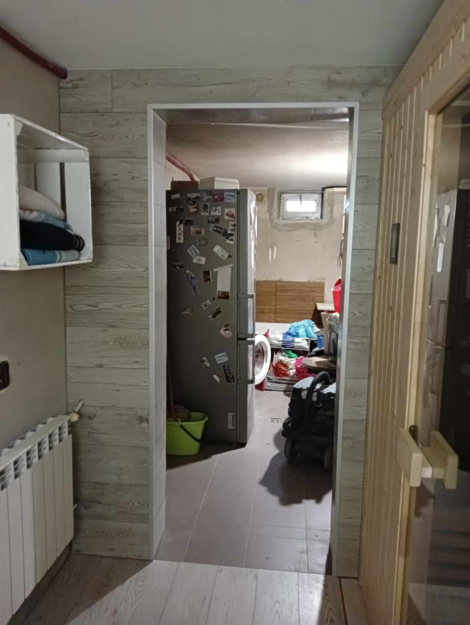 Widok przez futrynę na remontowane pomieszczenie gospodarcze z widoczną pralką i lodówką obklejoną magnesami, ściany wykończone panelami imitującymi drewno.