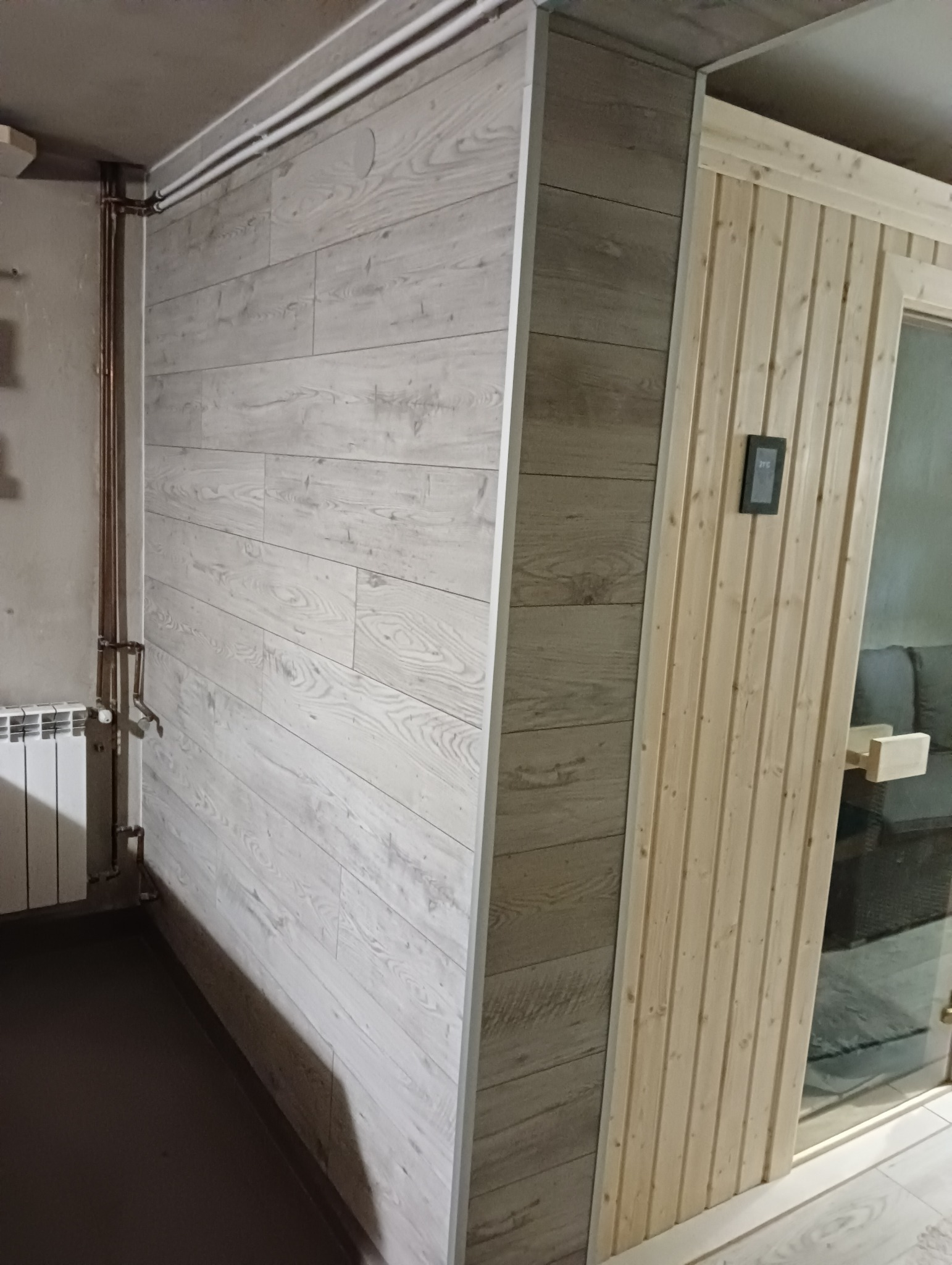 Wykończone wnętrze z panelami ściennymi imitującymi drewno i fragmentem sauny z drewnianymi panelami pionowymi oraz szklanymi drzwiami.