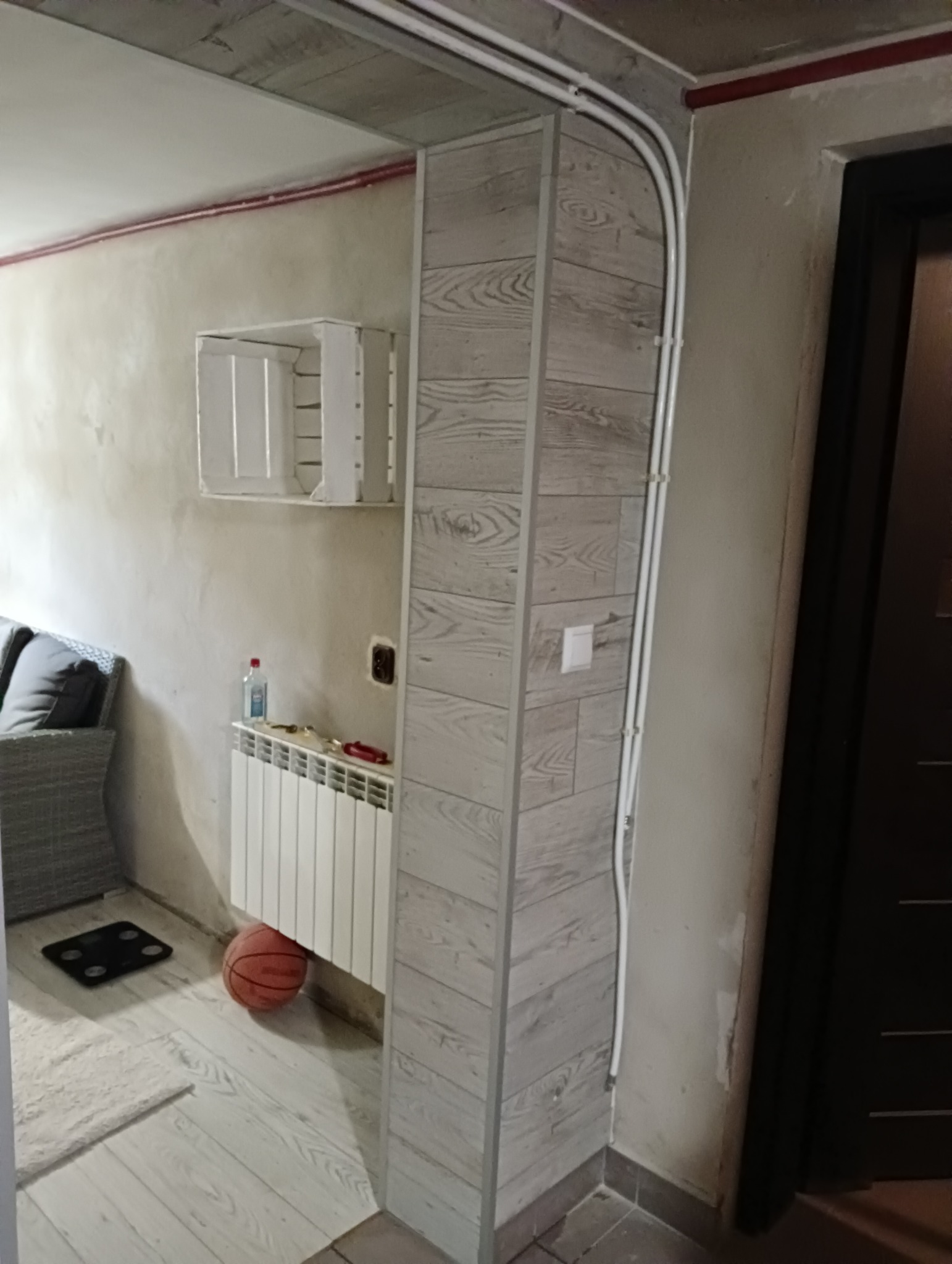 Wykończone wejście do pomieszczenia z obudowaną kolumną imitującą drewno, widoczne rury instalacyjne na ścianie, grzejnik, w tle sofa i biała półka, na podłodze waga łazienkowa i piłka do koszykówki.