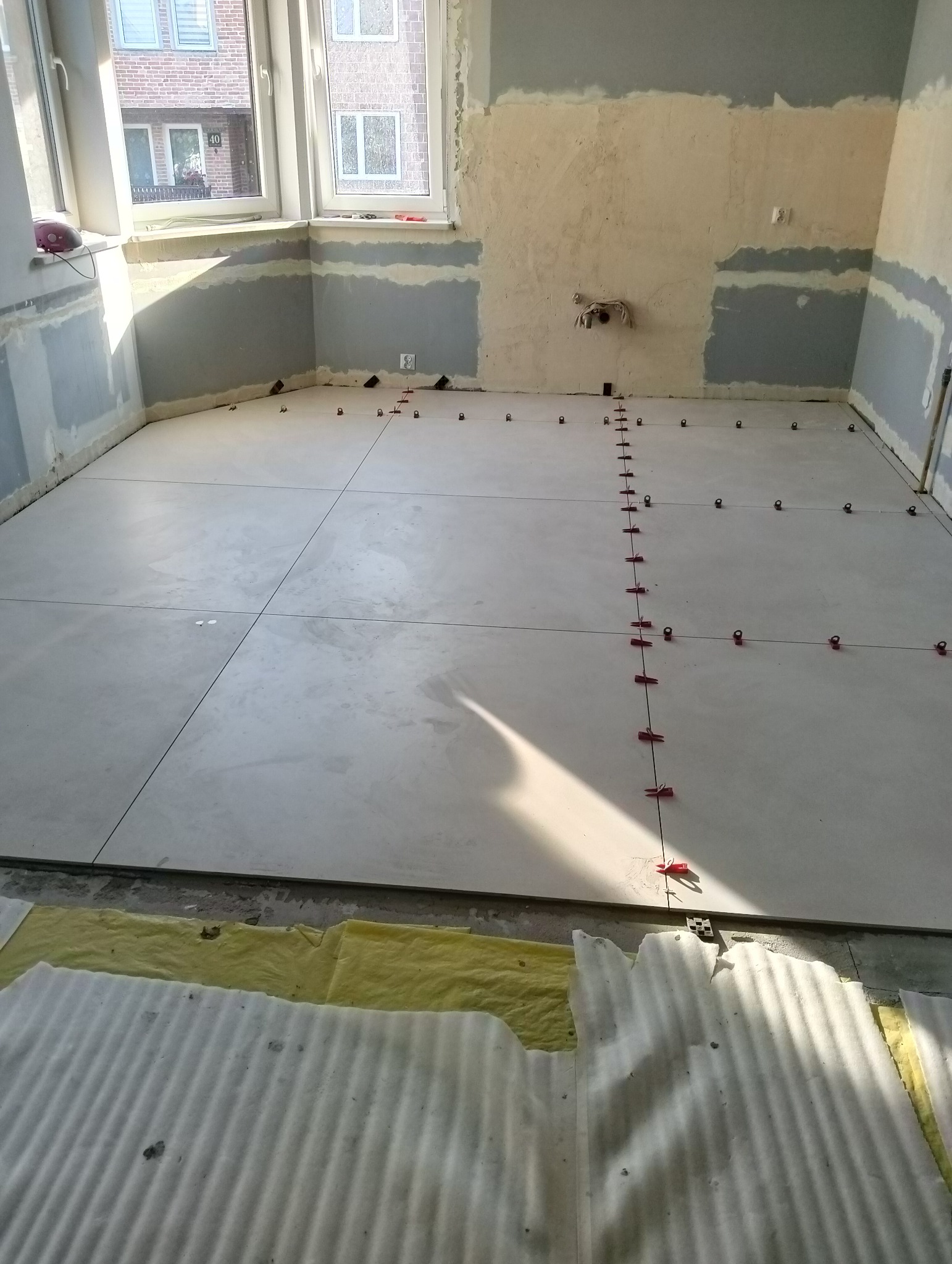 Układanie jasnych, kwadratowych płytek podłogowych w pokoju z przygotowanymi ścianami, widoczne krzyżyki dystansowe i materiał izolacyjny przy ścianie.