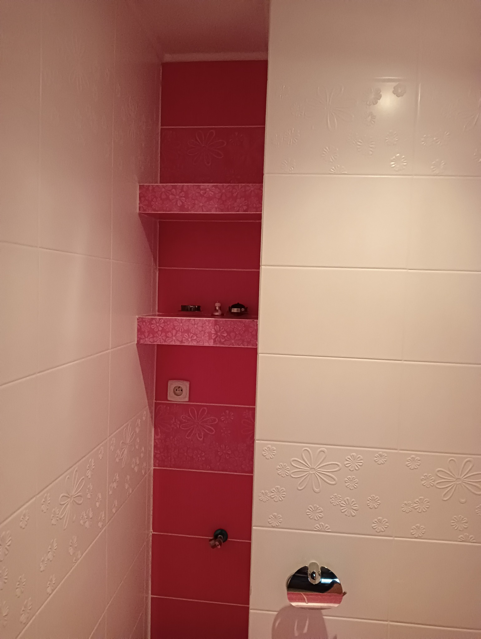 Wykończona łazienka z czerwono-białymi płytkami z motywem kwiatowym, wnęka z półkami wyłożona czerwonymi płytkami z kwiatowym wzorem, na półkach drobne przedmioty dekoracyjne, widoczne gniazdko...