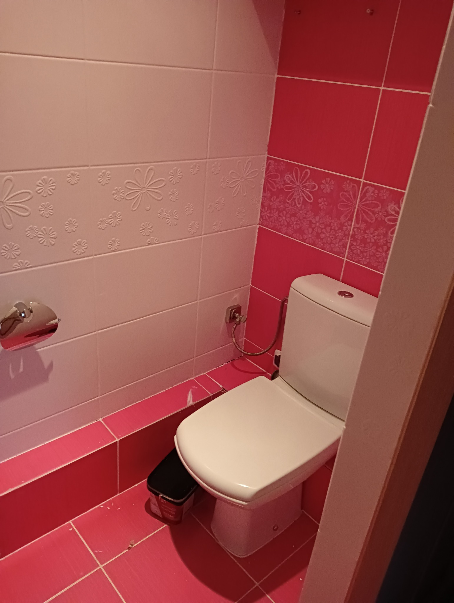 Nowoczesna toaleta z białą ceramiką, różowe płytki z kwiatowym wzorem, pojemnik na farbę obok muszli klozetowej, widoczny fragment futryny drzwi.