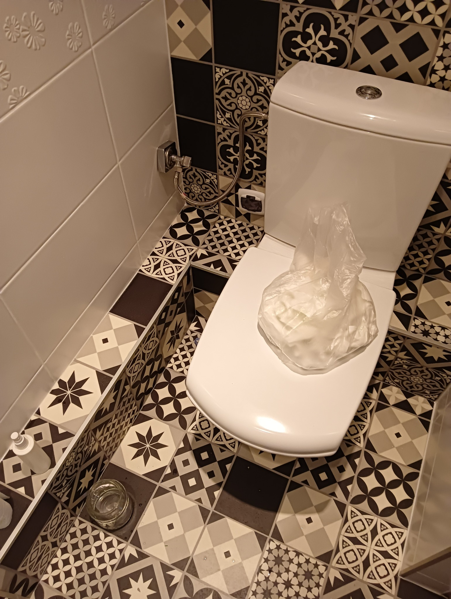 Biała toaleta z deską w łazience wyłożonej wzorzystymi, czarno-białymi płytkami. Na desce leży zapakowany w folię, nowy zestaw akcesoriów toaletowych. Widoczny fragment ściany z białymi płytkami...