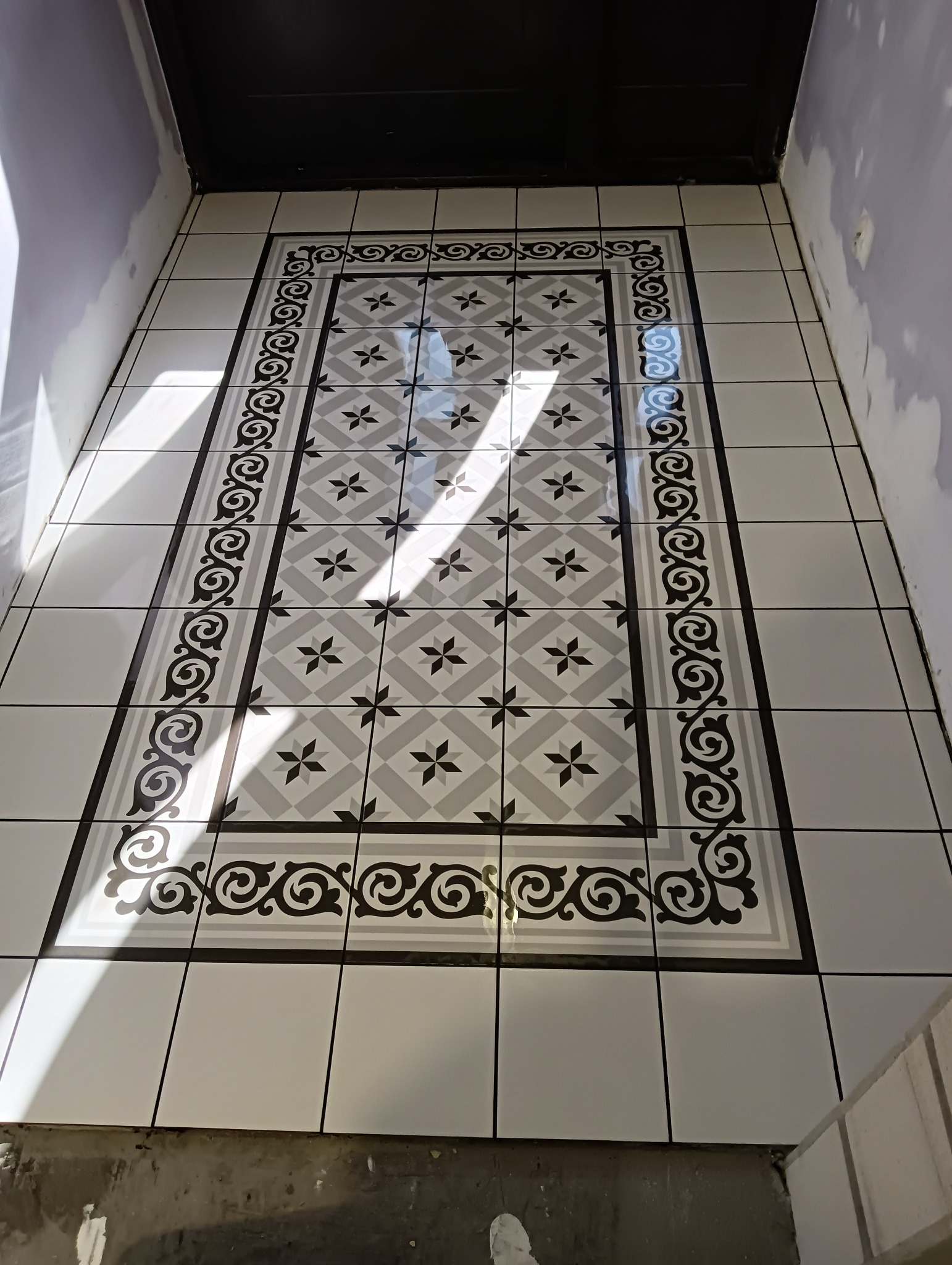 Wzorzysta podłoga w wiatrołapie, ułożona z białych płytek z szarym wzorem w romby i czarnym obramowaniem z ornamentami, widoczne fragmenty ścian przed wykończeniem.