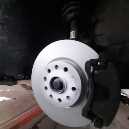 AUTO SERWIS KUBIAK PIOTR KUBIAK - Nowa tarcza hamulcowa zamontowana w samochodzie, widoczny zacisk hamulcowy i zawieszenie, w tle podnośnik samochodowy.