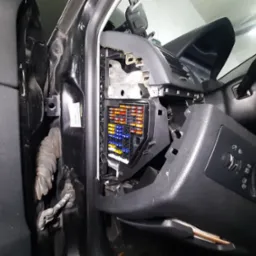 Otwarty panel bezpieczników w samochodzie z widocznymi kolorowymi bezpiecznikami i elementami deski rozdzielczej.