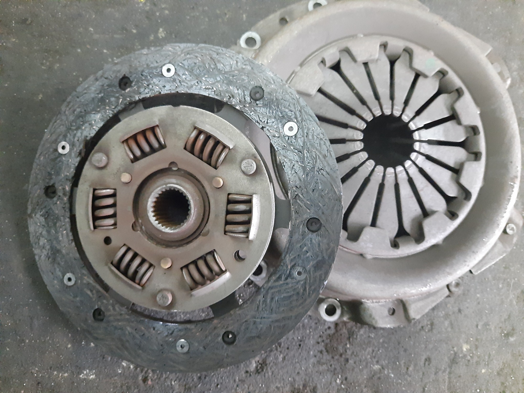 Dwie tarcze sprzęgła samochodowego, jedna nowa i czysta, druga zużyta z widocznymi śladami eksploatacji, leżące na szarym, betonowym podłożu.
