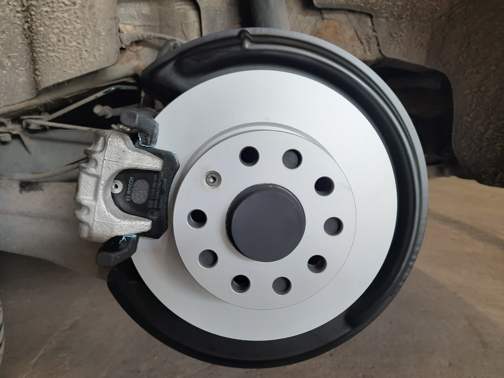 Nowa, srebrna tarcza hamulcowa z czarnym środkiem i zaciskiem hamulcowym marki Bosch, zamontowana w samochodzie. Widoczne ślady użytkowania na elementach zawieszenia.
