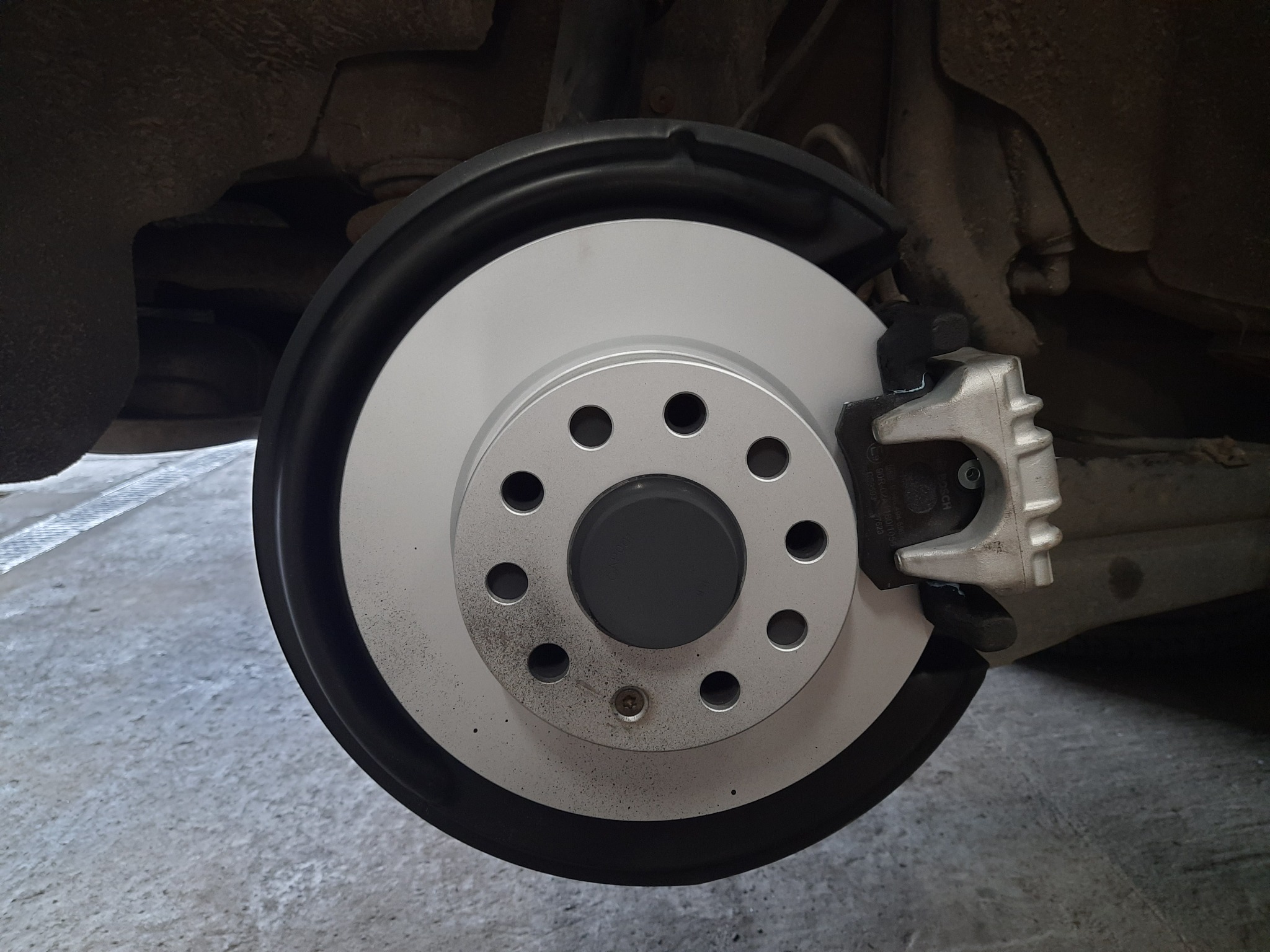 Widok z bliska nowego, srebrnego tarczy hamulcowej z zaciskiem zamontowanej w samochodzie, widoczne ślady montażu.