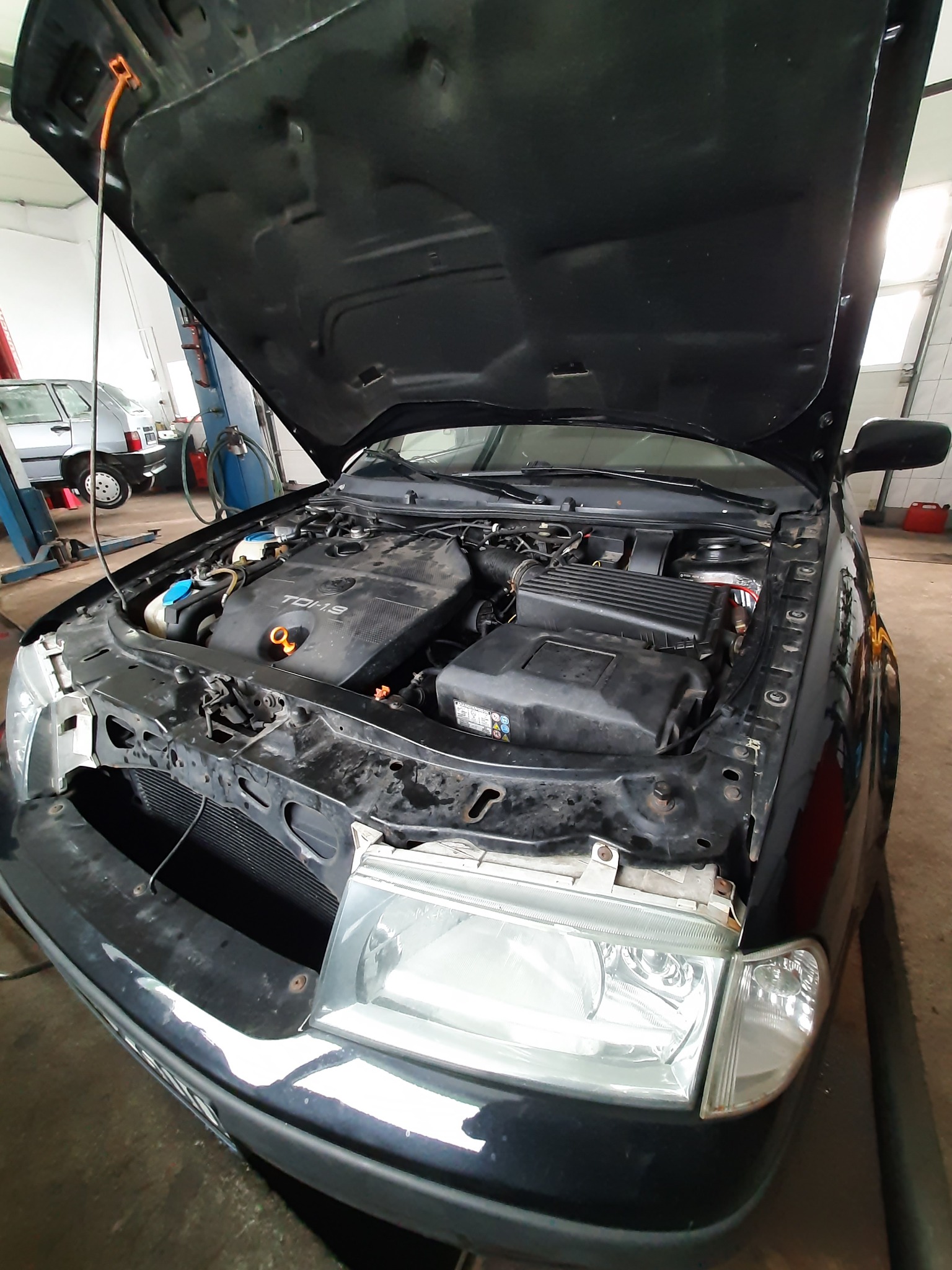 Czarny samochód z otwartą maską w warsztacie, widoczny silnik TDI 1.9 oraz reflektor. W tle podnośnik kolumnowy i inny pojazd.