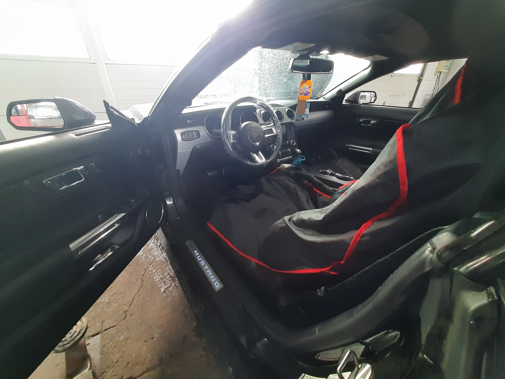 Wnętrze samochodu Ford Mustang z założonym pokrowcem ochronnym na fotelu i folią na kierownicy, widoczne detale deski rozdzielczej i oświetlenie progu.