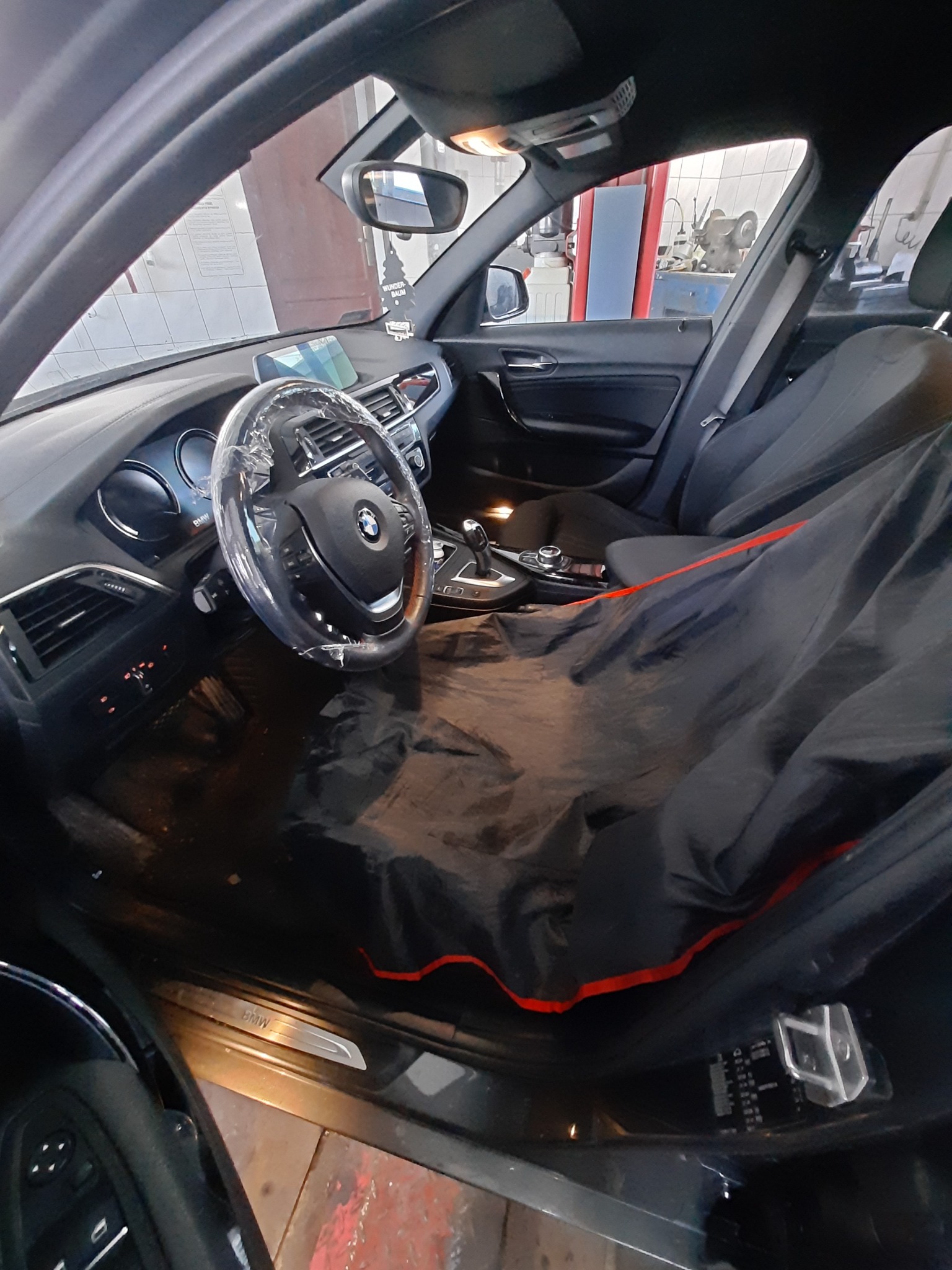 Wnętrze samochodu BMW, zabezpieczone folią na kierownicy i pokrowcem ochronnym na siedzeniu, widoczne wyposażenie warsztatu w tle.