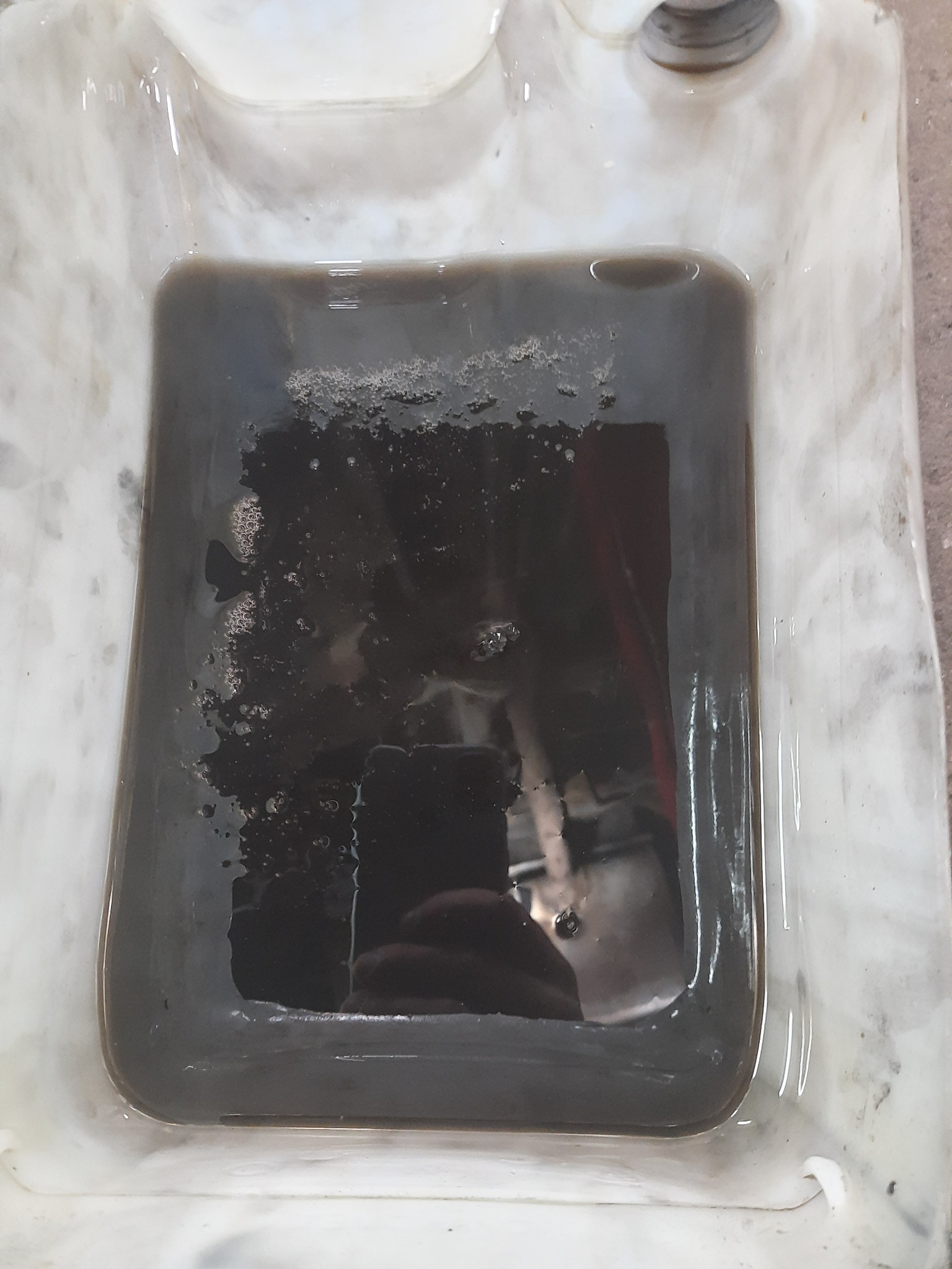 Zużyty, ciemny olej silnikowy w plastikowym pojemniku, widoczne pęcherzyki powietrza i osad na dnie.