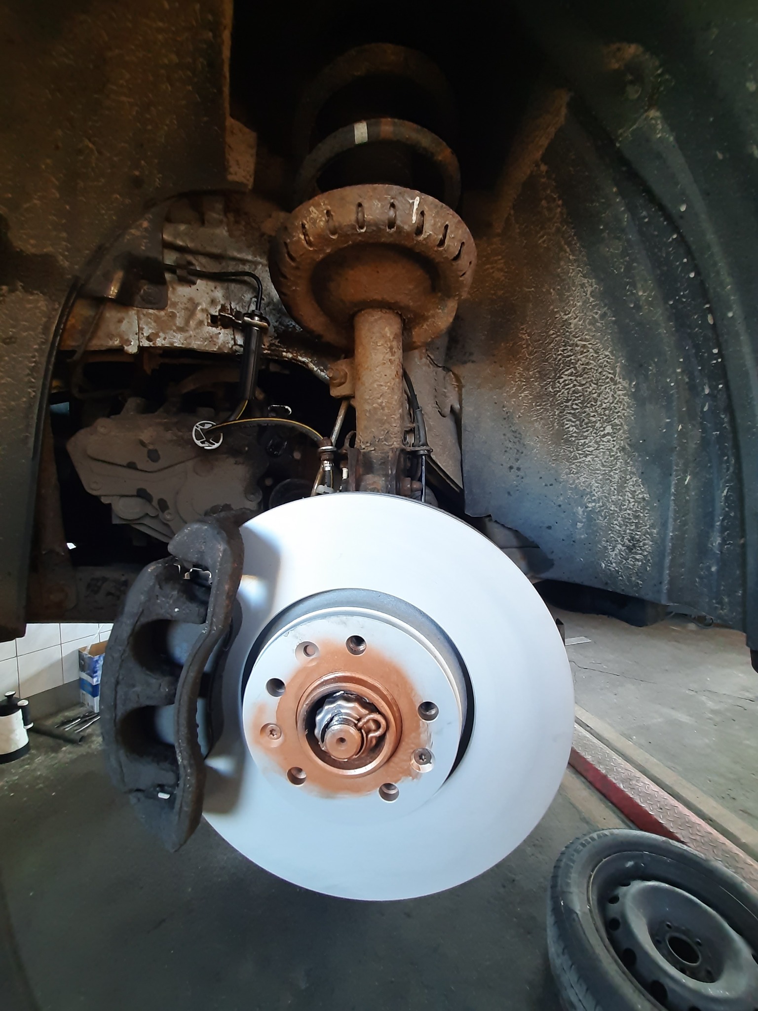 Nowa tarcza hamulcowa zamontowana w samochodzie, widoczne zaciski hamulcowe, amortyzator i elementy zawieszenia, w tle fragment opony.