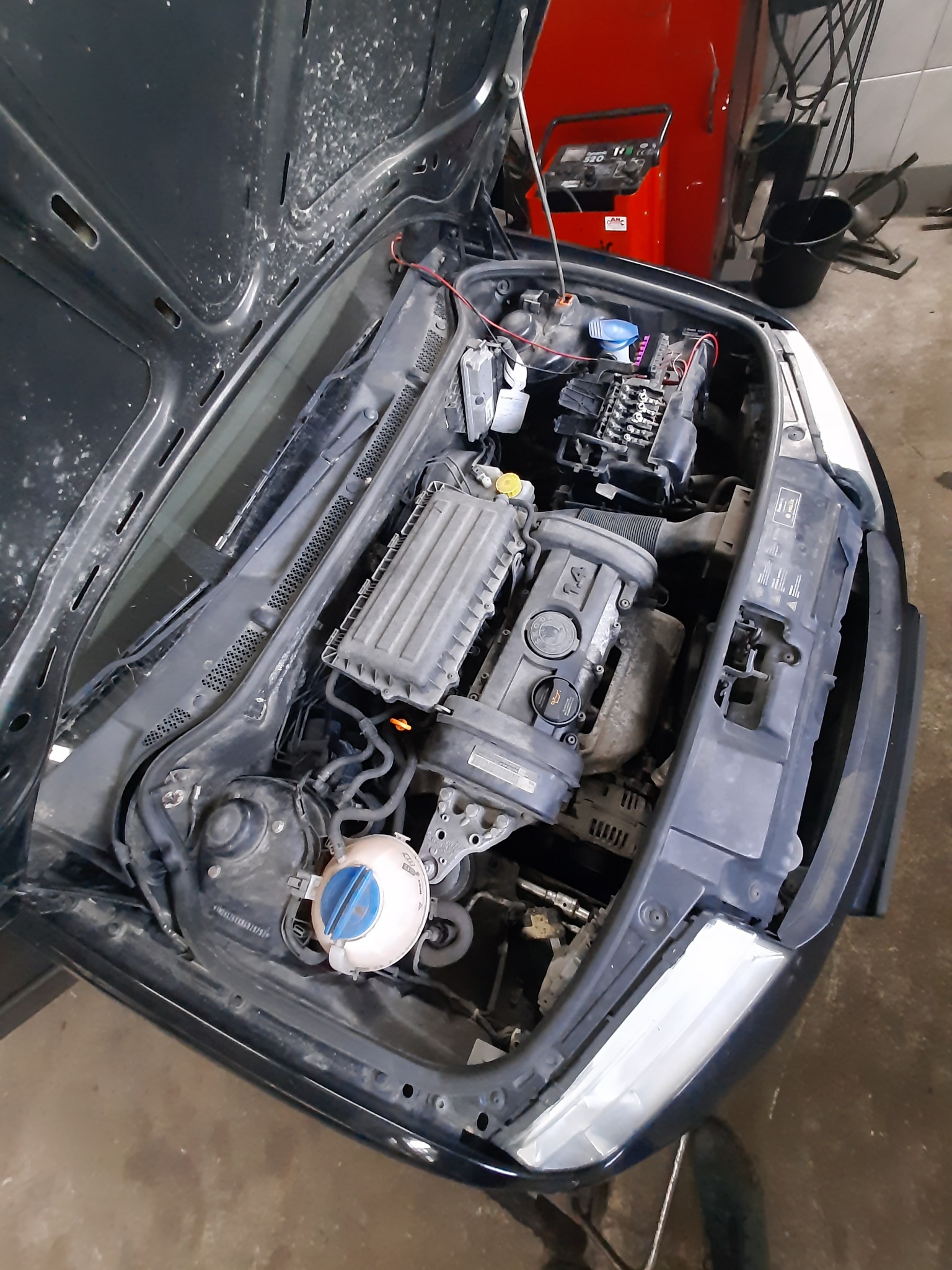 Otwarta maska samochodu z widocznym silnikiem i elementami osprzętu, w tle czerwone urządzenie diagnostyczne.