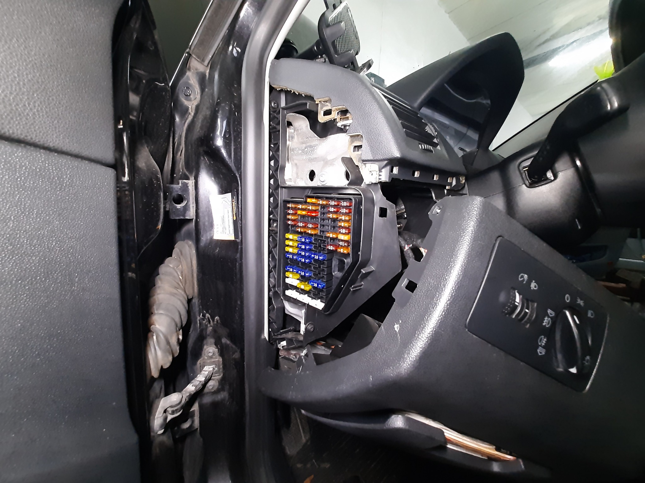 Otwarty panel bezpieczników w samochodzie z widocznymi kolorowymi bezpiecznikami i elementami deski rozdzielczej.