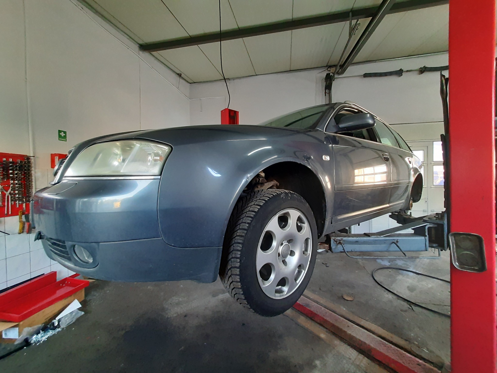 Szary samochód Audi A6 na podnośniku w warsztacie samochodowym, z widocznym kołem z oponą zimową i zdjętym drugim kołem, narzędzia na ścianie w tle.