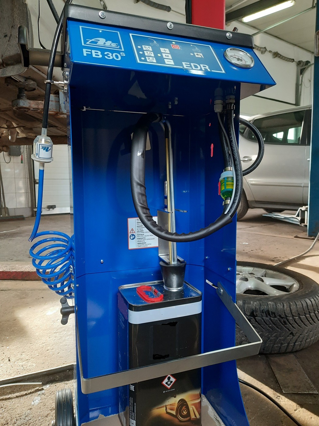 Niebieskie urządzenie ATE FB 30S EDR do wymiany płynu hamulcowego w warsztacie samochodowym, widoczny podnośnik z podwieszonym autem i opona w tle.