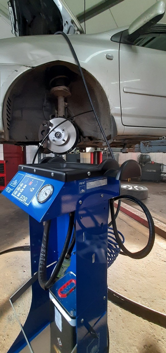 Samochód na podnośniku z otwartą maską, widoczny nowy element układu hamulcowego i niebieski automat do płukania układów hamulcowych ATE FB 30 EDR.