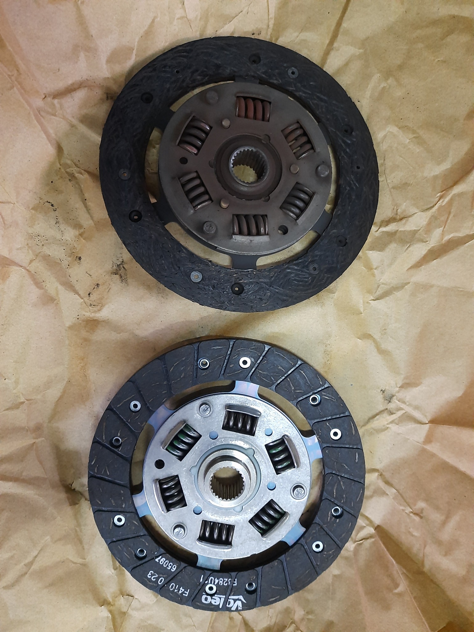 Dwie tarcze sprzęgłowe, używana i nowa, leżące na brązowym papierze. Górna tarcza jest zużyta, dolna nowa z widocznym logo Valeo.