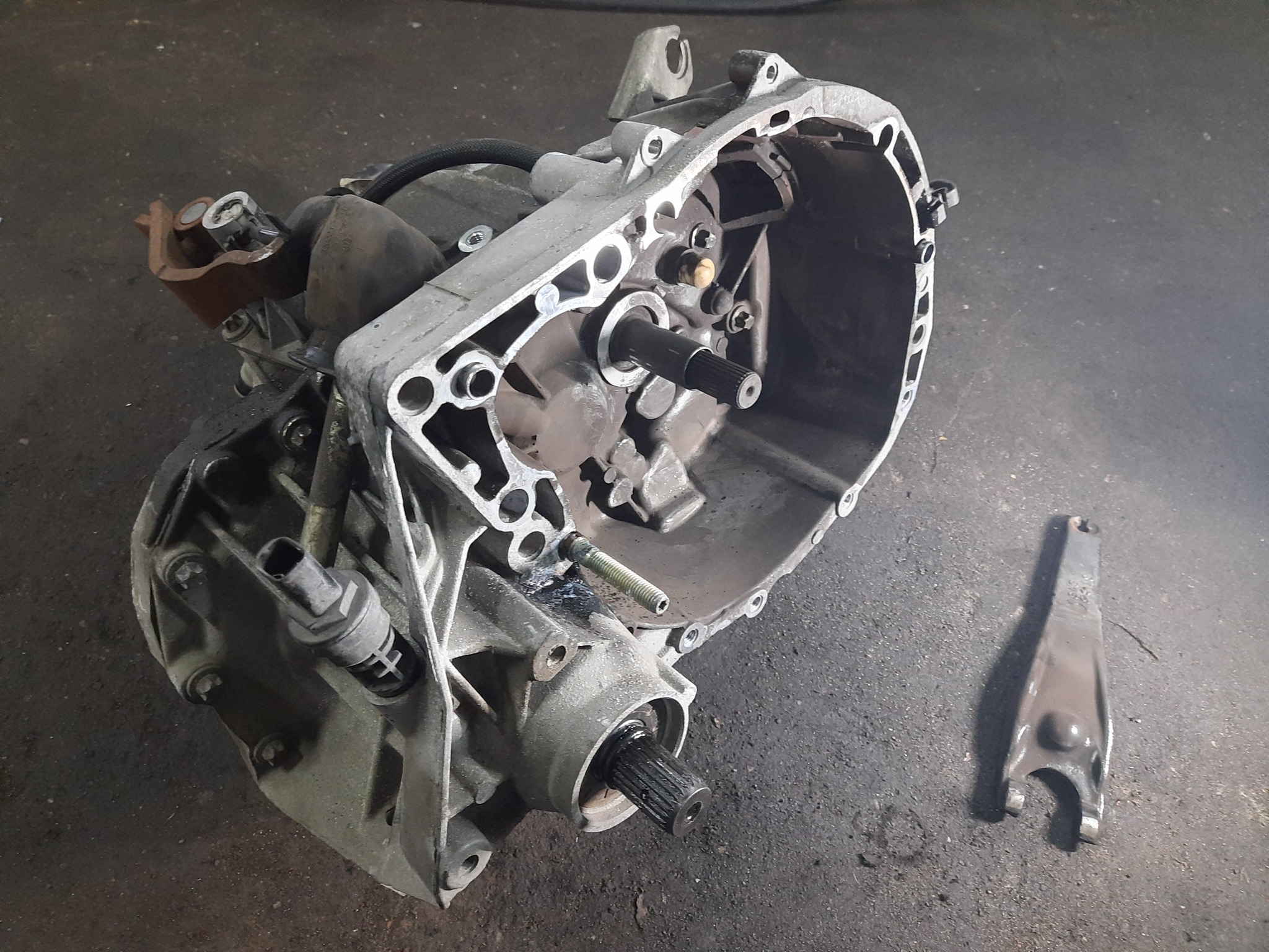 Zdjęcie skrzyni biegów samochodu w trakcie demontażu, widoczne elementy wewnętrzne i obudowa, obok leży widelec zmiany biegów na brudnej podłodze warsztatowej.