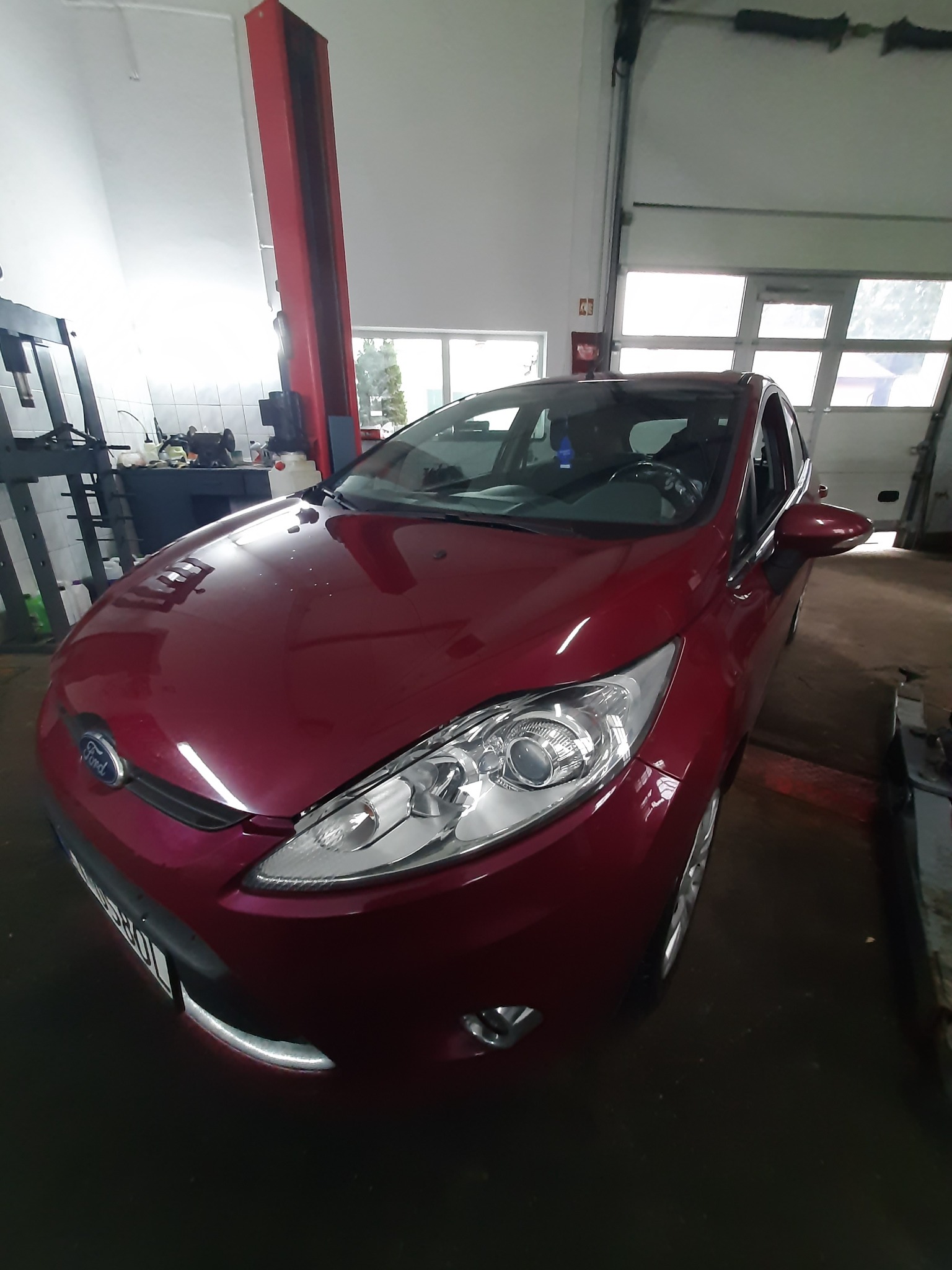 Bordowy Ford Fiesta w warsztacie samochodowym, widoczny podnośnik i narzędzia w tle.