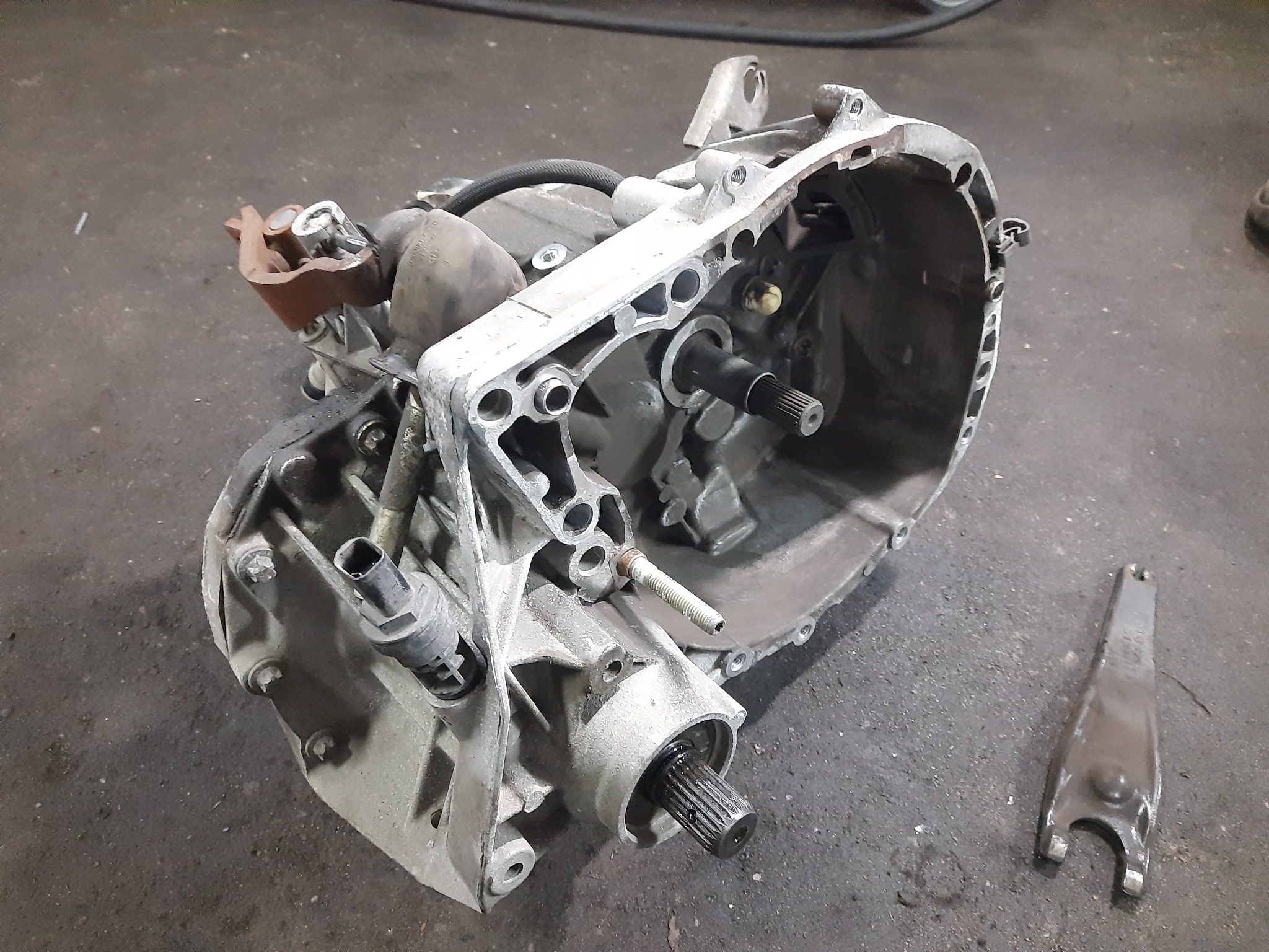 Otwarta skrzynia biegów samochodu, widoczne elementy wewnętrzne, obudowa aluminiowa, lewarek zmiany biegów obok, na brudnej podłodze warsztatu.