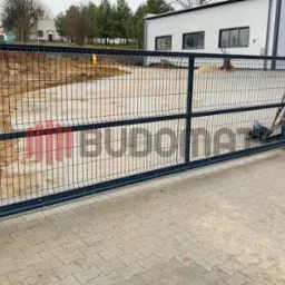 Przesuwna brama wjazdowa z paneli metalowych, widoczna na tle budynku przemysłowego z placem wyłożonym kostką brukową.